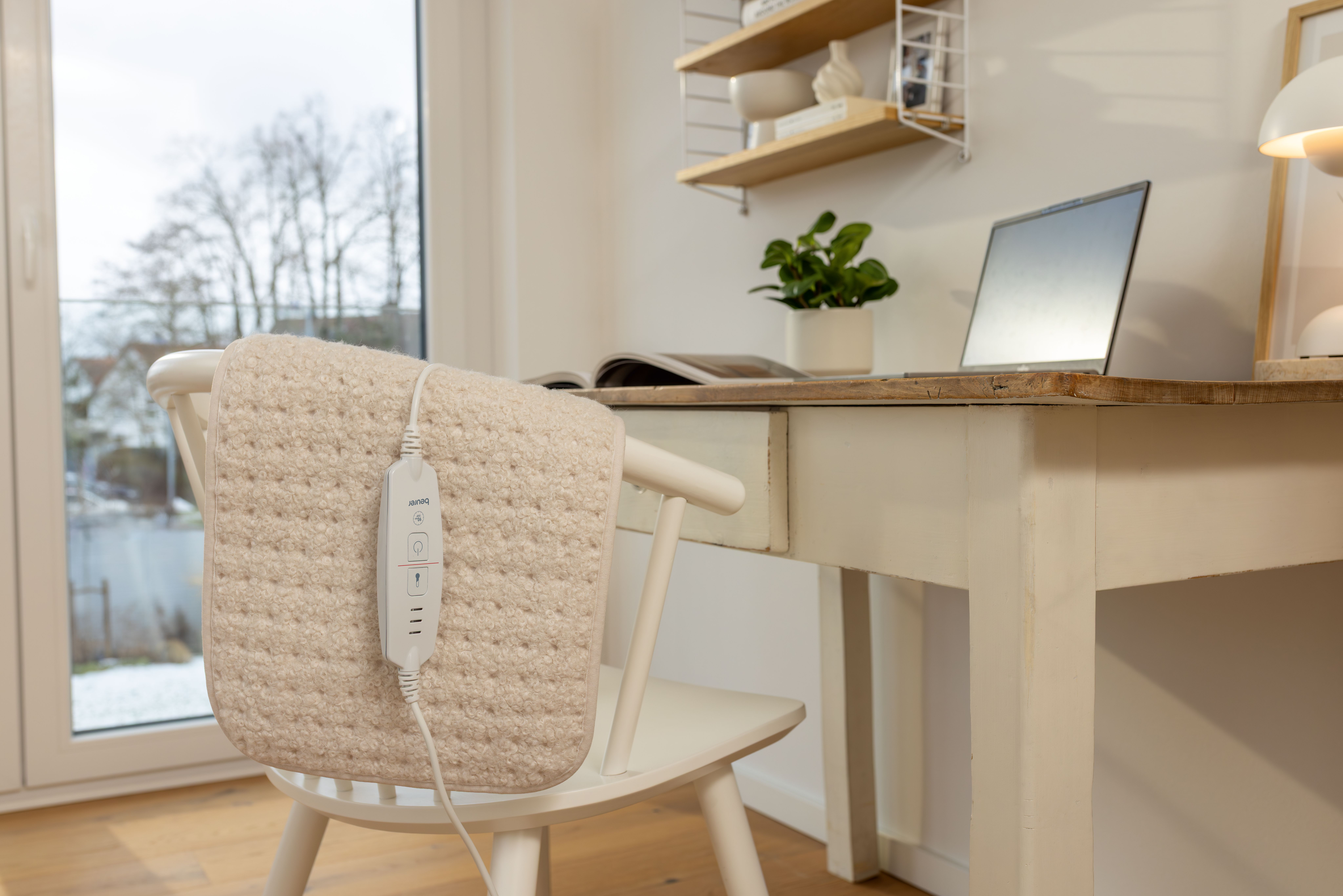 Ein beiger, flauschiger Heizkissen auf einem weißen Stuhl vor einem Holztisch mit Laptop und Pflanze.
