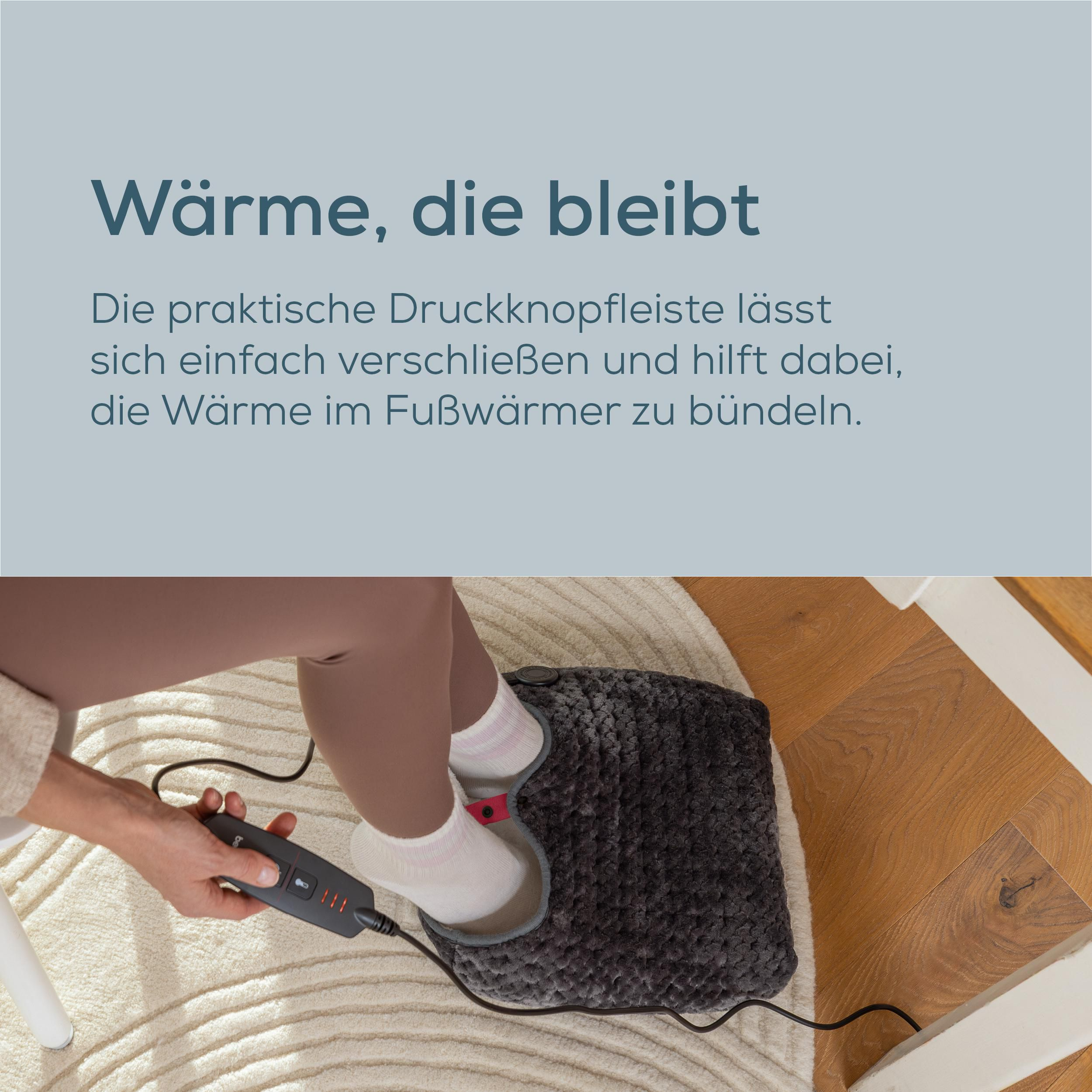 Eine Person sitzt auf einem Stuhl, wärmt ihre Füße in einem grauen elektrischen Fußwärmer und bedient das Steuergerät.