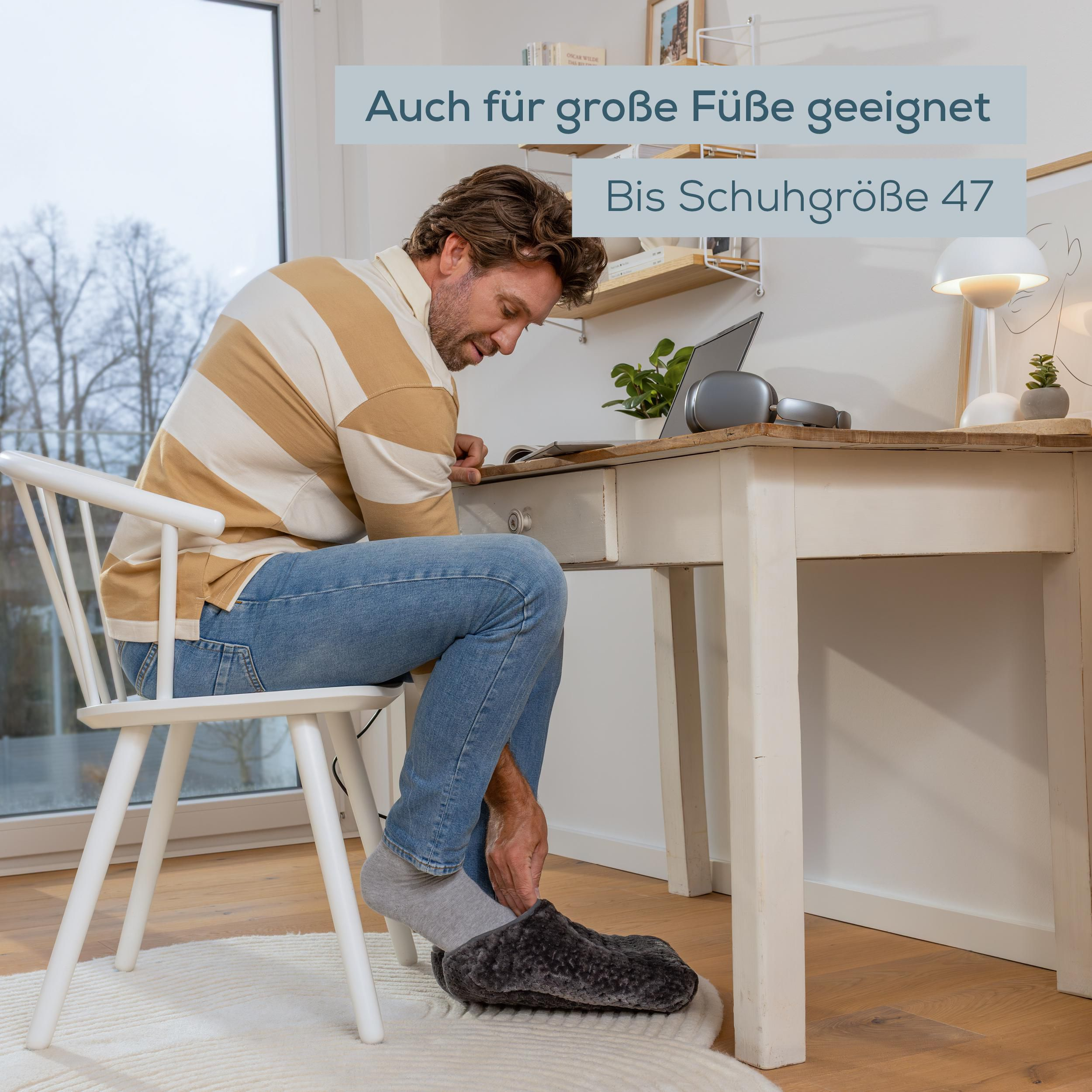 Ein Mann sitzt an einem Holztisch in einem hellen Raum und zieht große, graue Fußwärmer an.