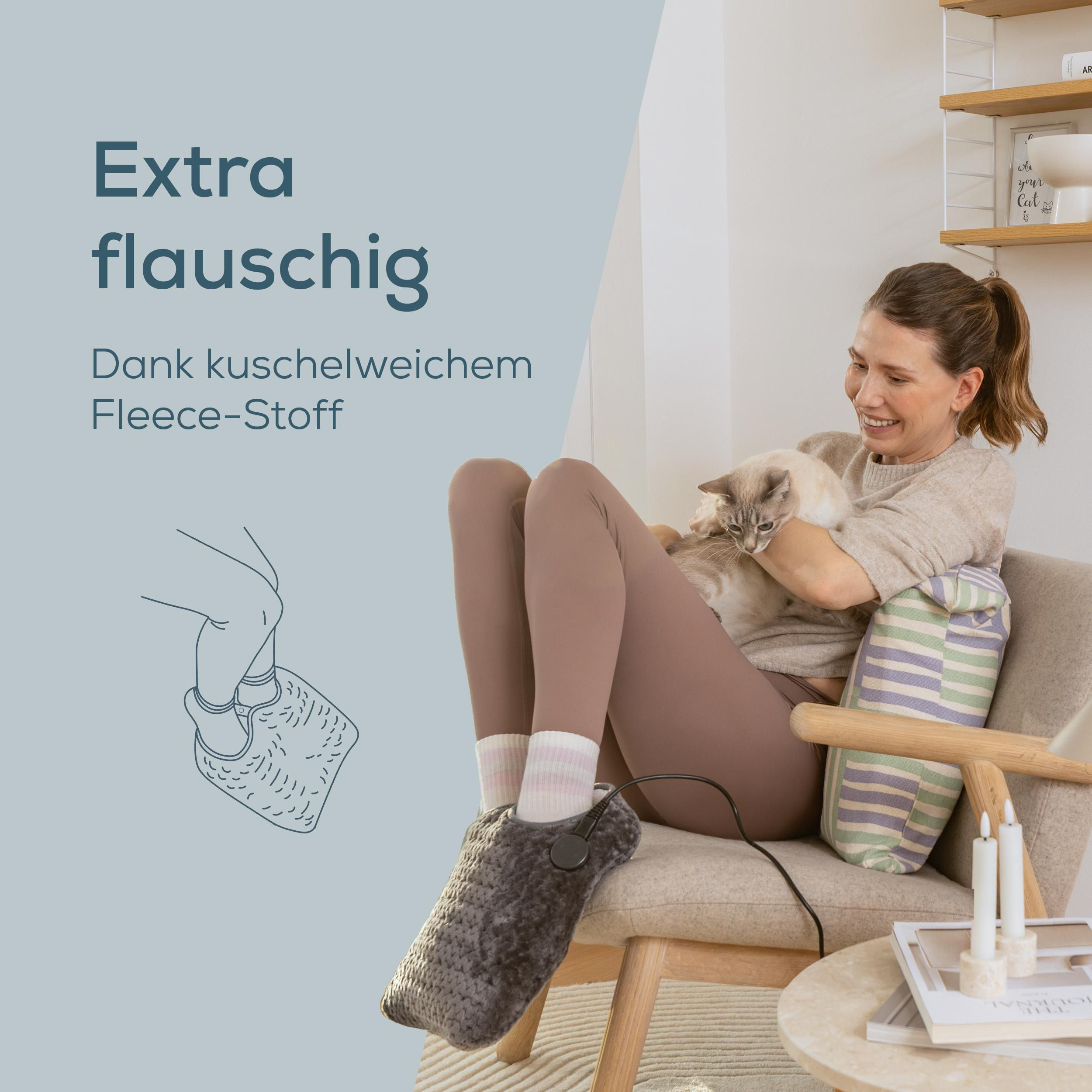 Eine Frau sitzt auf einem Sessel, hält eine Katze im Arm und trägt einen grauen, beheizbaren Fußwärmer.