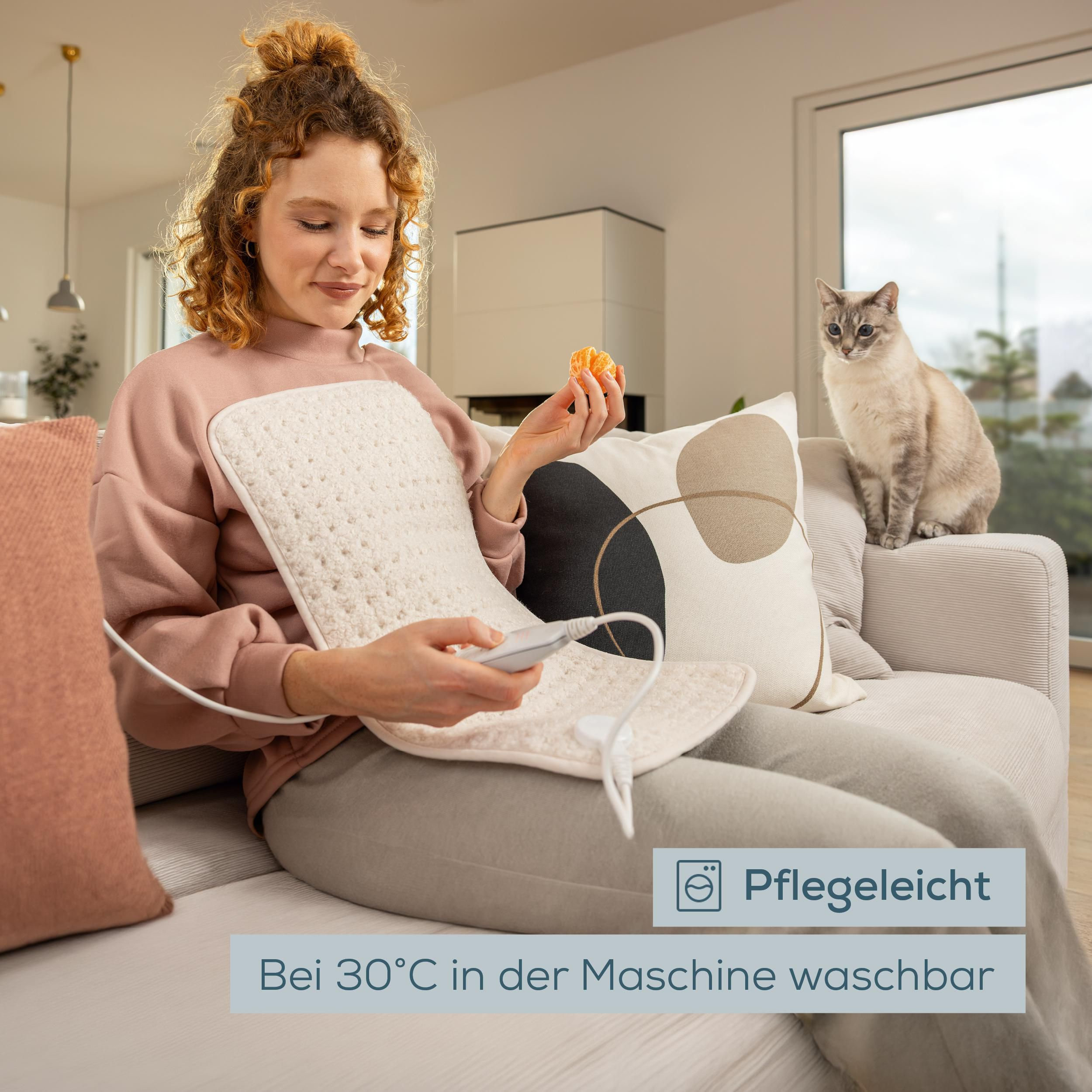 Eine Frau sitzt auf einem Sofa, hält ein Heizkissen und eine Mandarine, während eine Katze zusieht.