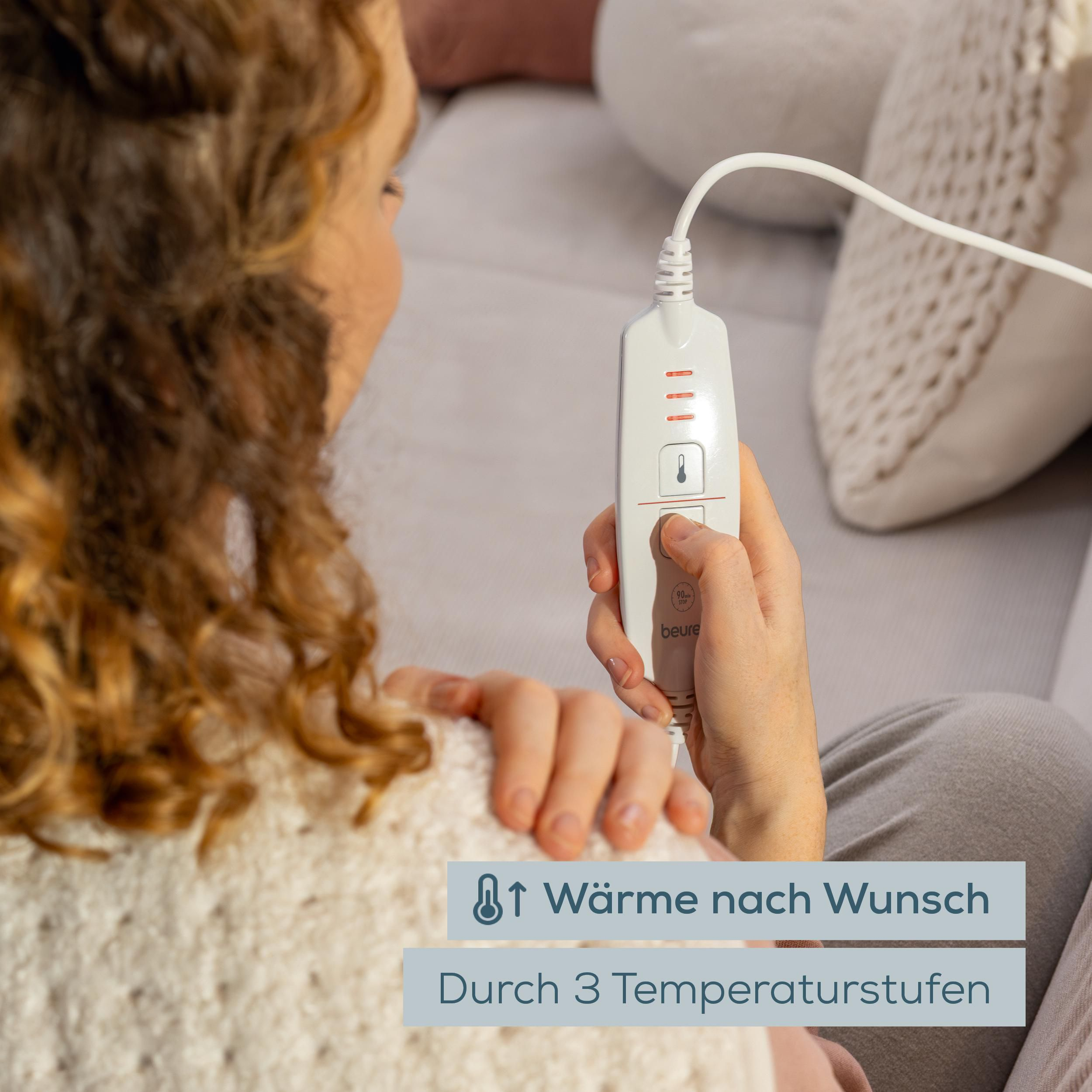 Frau sitzt auf dem Sofa und hält eine weiße Fernbedienung für ein Heizkissen mit drei Temperaturstufen.