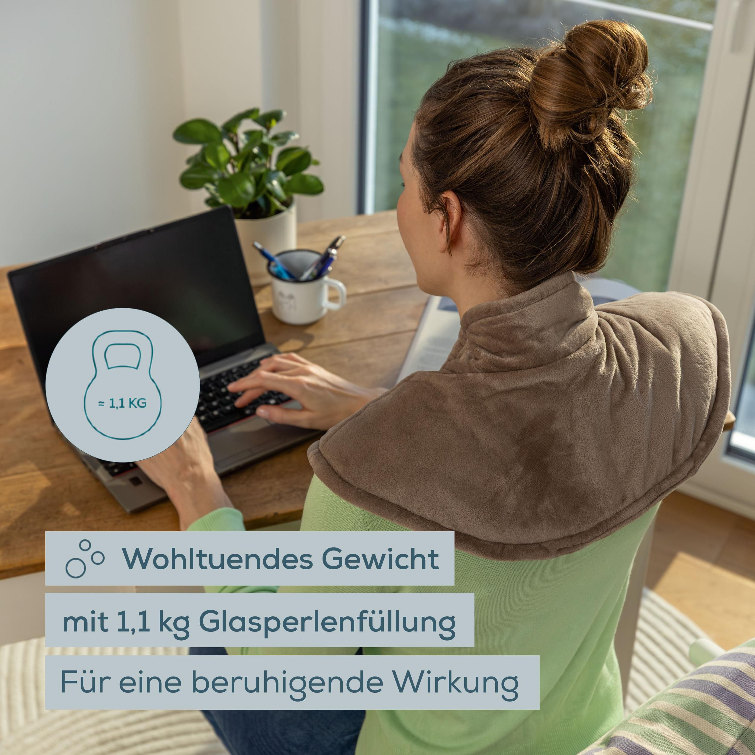 Eine Frau sitzt mit einer braunen Gewichts-Nackenkissen-Heizdecke an einem Laptop am Holztisch.