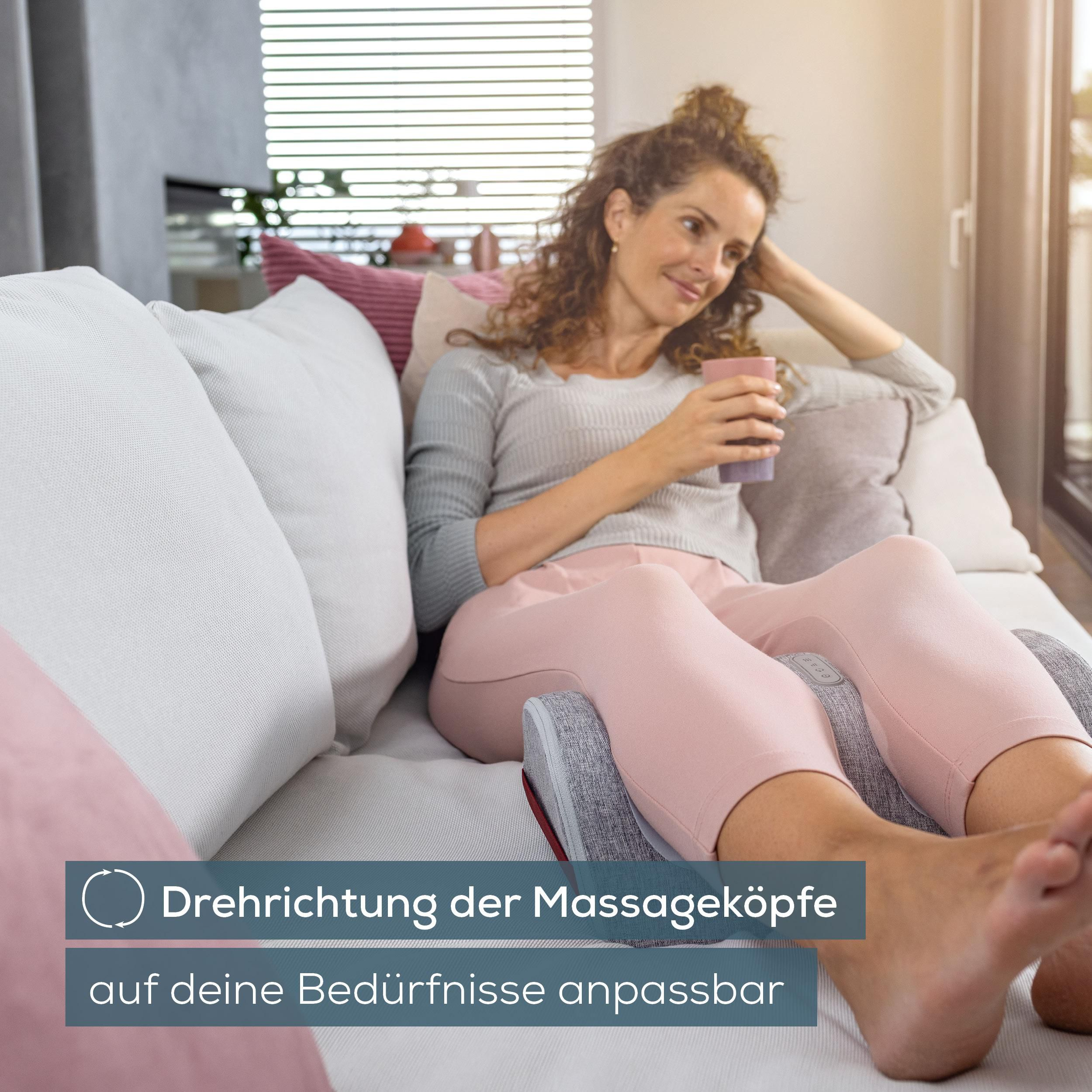 Eine Frau entspannt auf einem Sofa, hält eine Tasse und ruht ihre Beine auf einem Massagegerät aus.