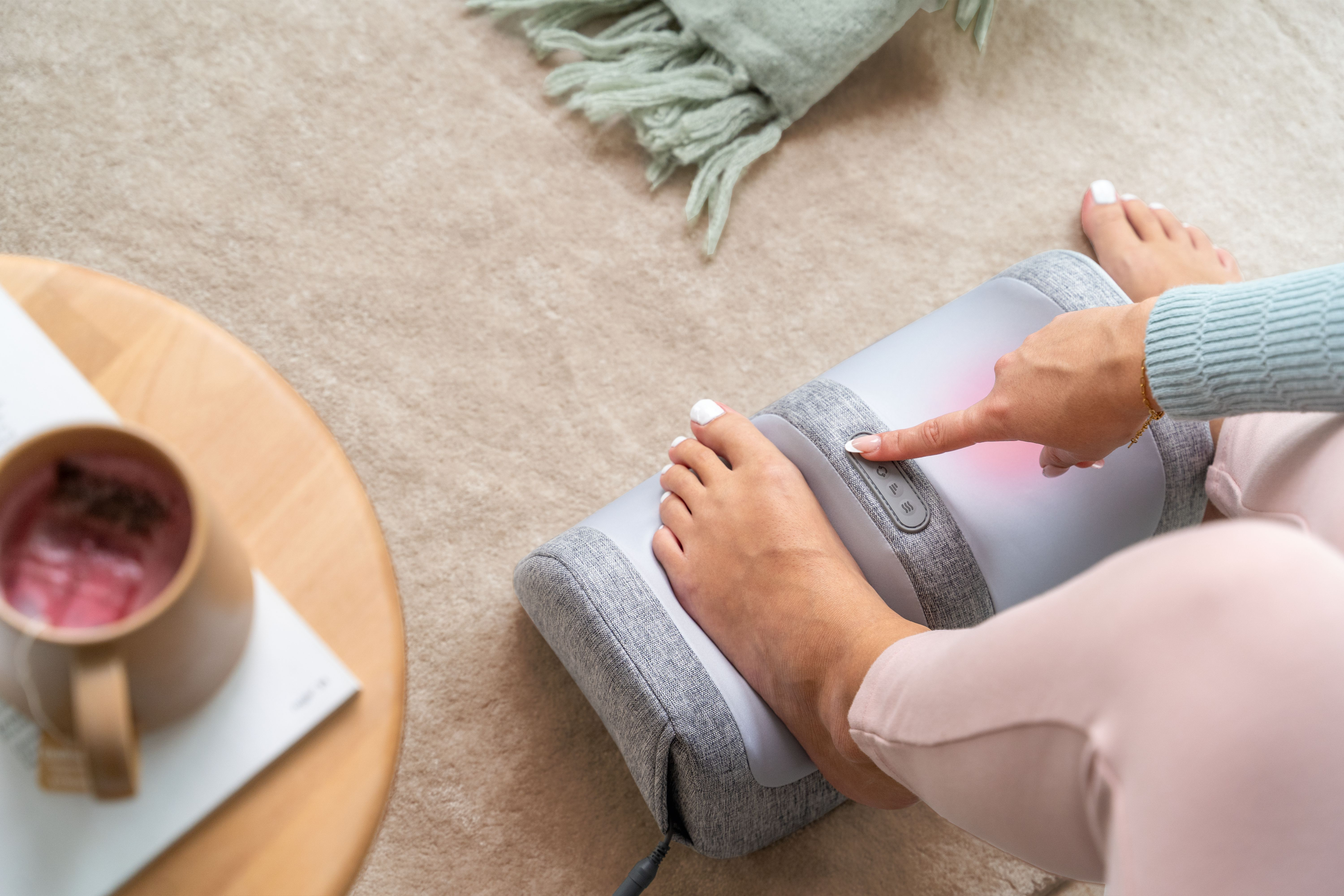 Eine Frau bedient ein graues Fußmassagegerät auf einem Teppich, neben einem Holztisch mit einer Tasse.