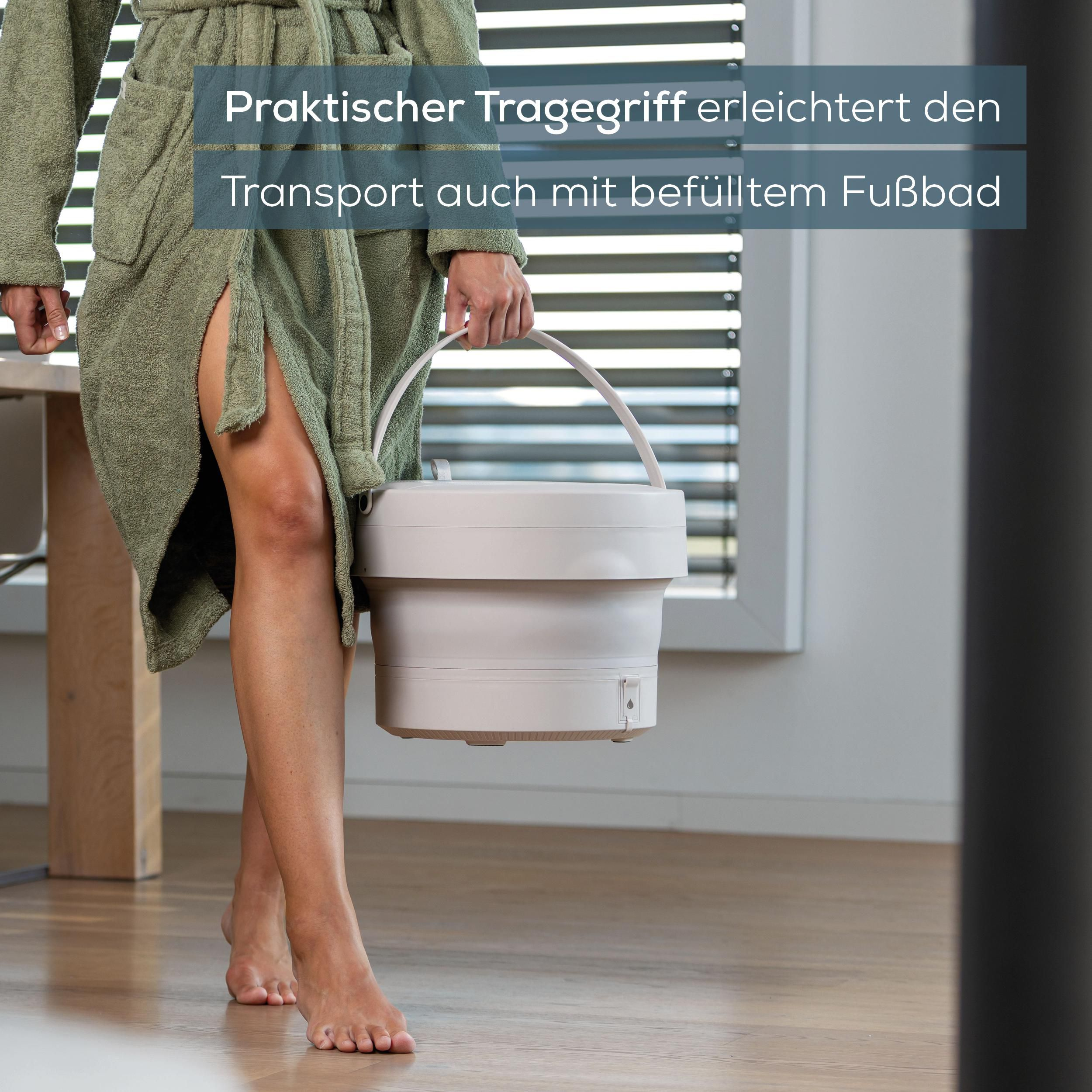Eine Person im grünen Bademantel trägt ein beiges, faltbares Fußbad mit praktischem Griff.