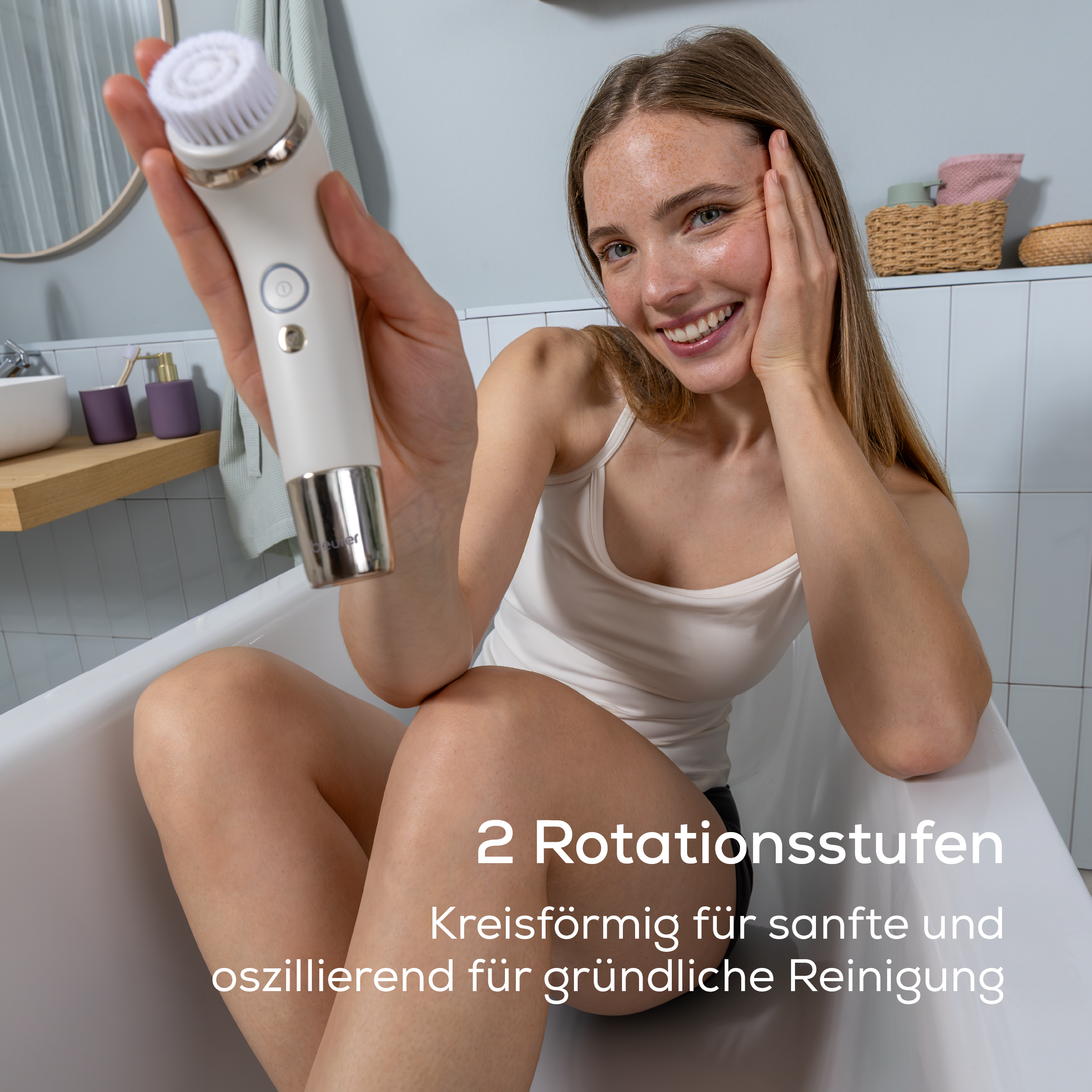 Eine lächelnde Frau sitzt in einer Badewanne und hält eine weiße Gesichtsreinigungsbürste in die Kamera.