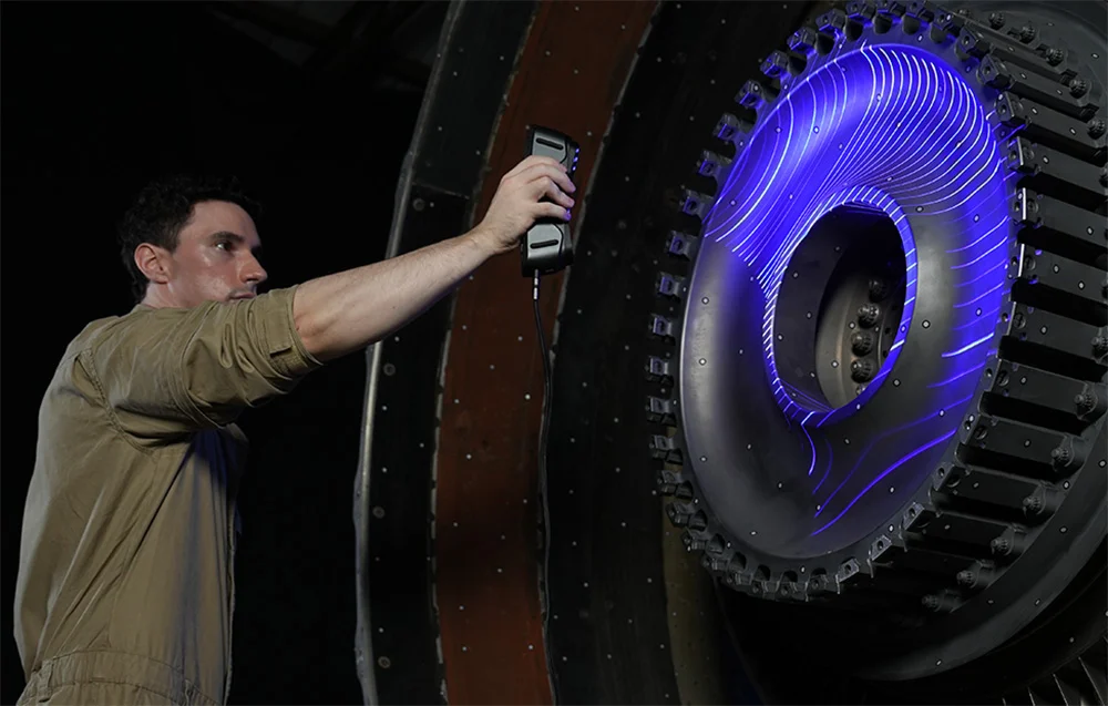 Egy dolgozó kézi szkennerrel kék lézerfényt vetít egy nagy ipari turbina alkatrészre.