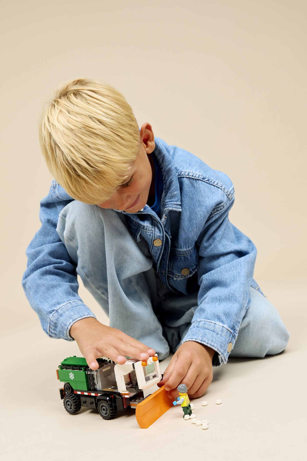 Un enfant blond en jean joue avec un véhicule Lego et une petite figurine sur fond clair.