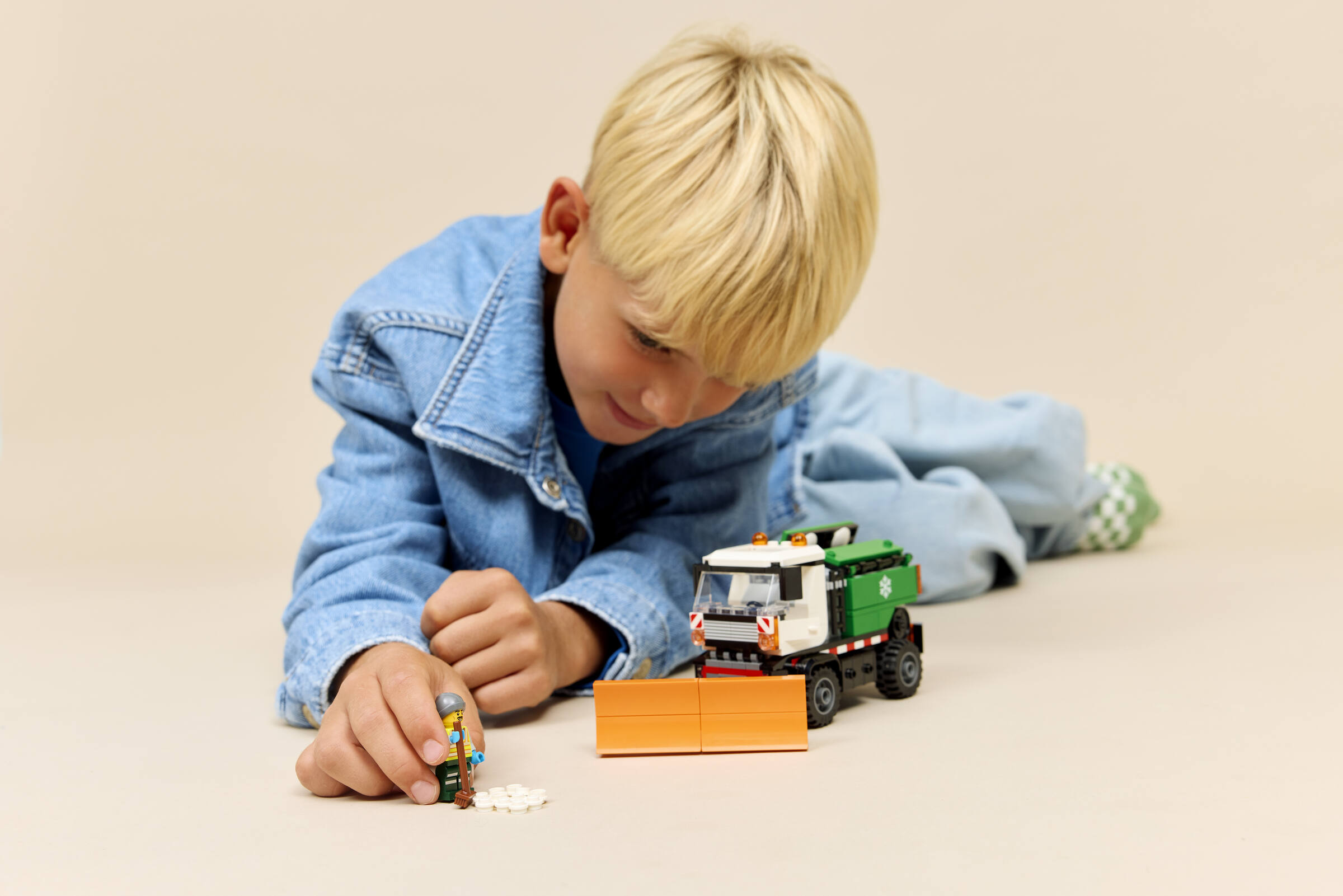 Un enfant blond en veste en jean joue par terre avec un camion Lego et une figurine.