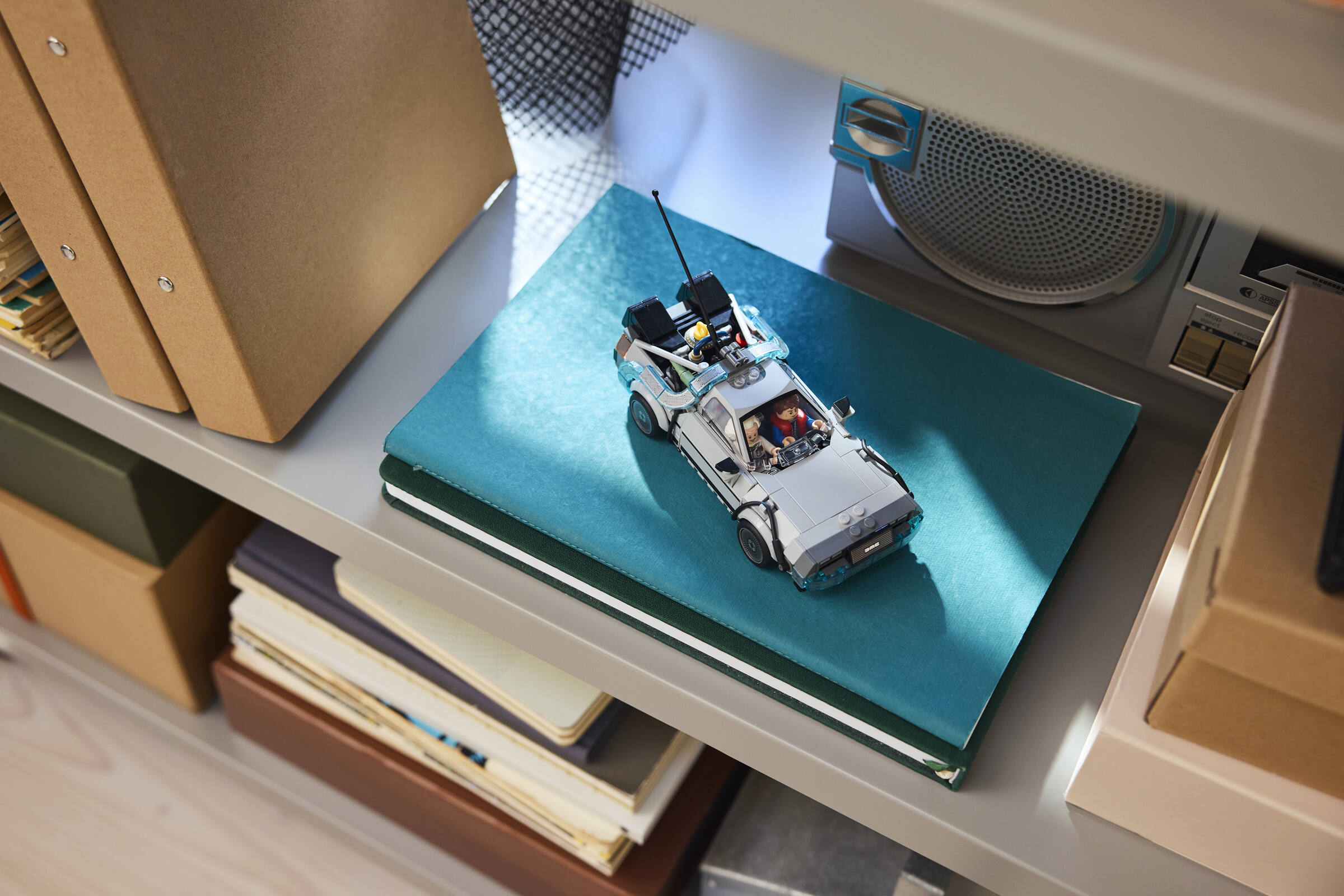 Ein Lego DeLorean Modellauto liegt auf einem türkisblauen Buch im Regal neben anderen Büchern und einem Radio.