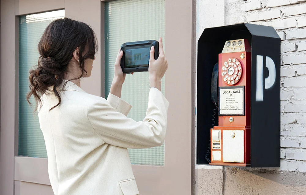 Egy fehér kabátos nő tablettel fényképez egy piros fali nyilvános telefondoboz.