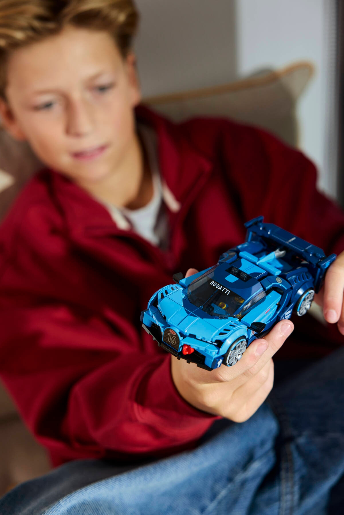 Un garçon en sweat à capuche rouge tient une voiture jouet Lego Bugatti bleue.