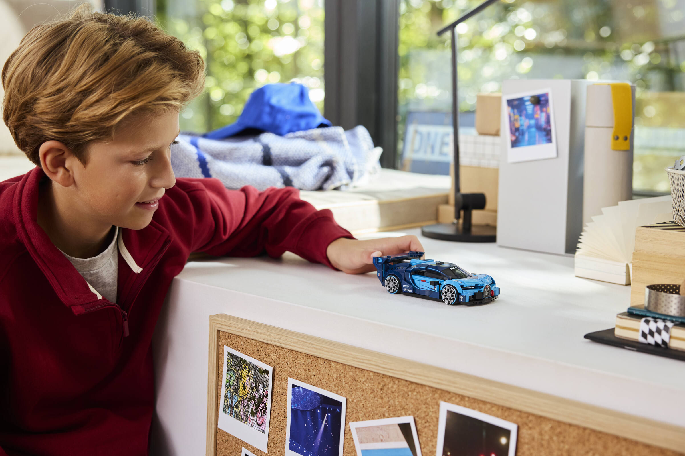 Un enfant blond portant un pull rouge joue avec une voiture LEGO bleue sur un bureau.