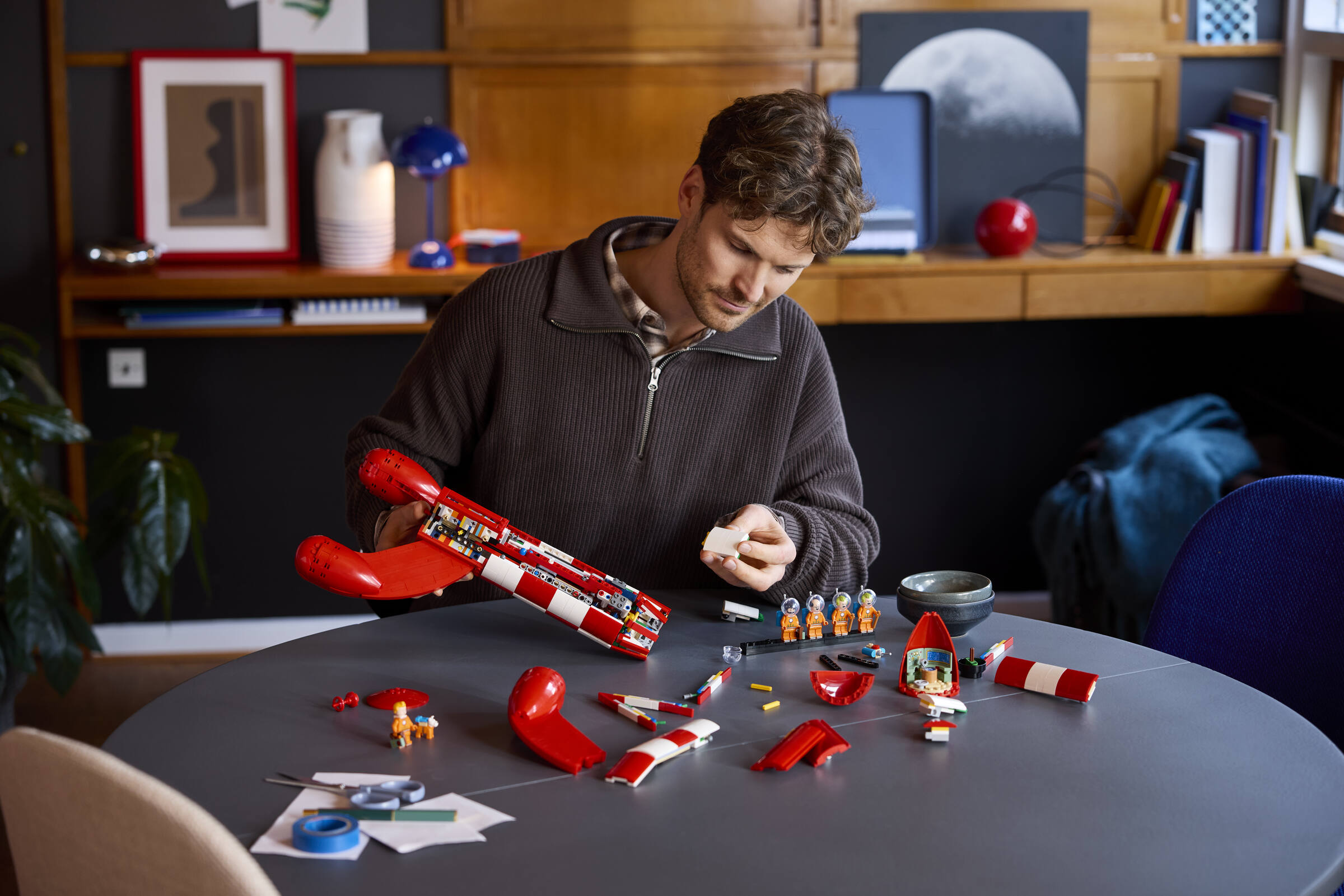 Un homme assemble un vaisseau spatial Lego rouge sur une table ronde, entouré de pièces et de figurines.