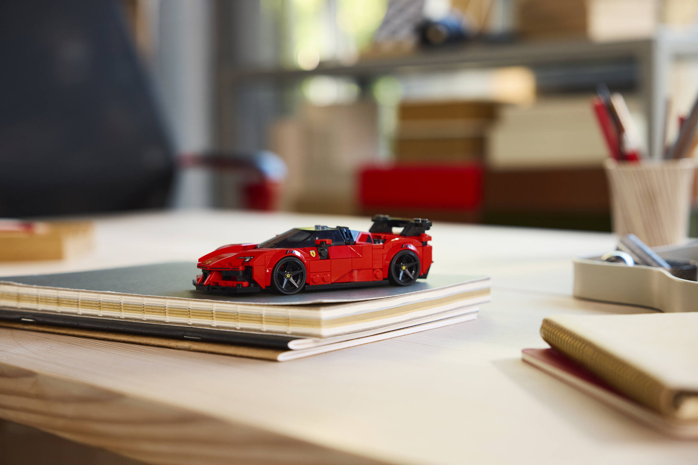 Ein rotes Lego-Spielzeugauto sitzt auf einem Notizbuch auf einem Holztisch, im Hintergrund ein Büroraum.