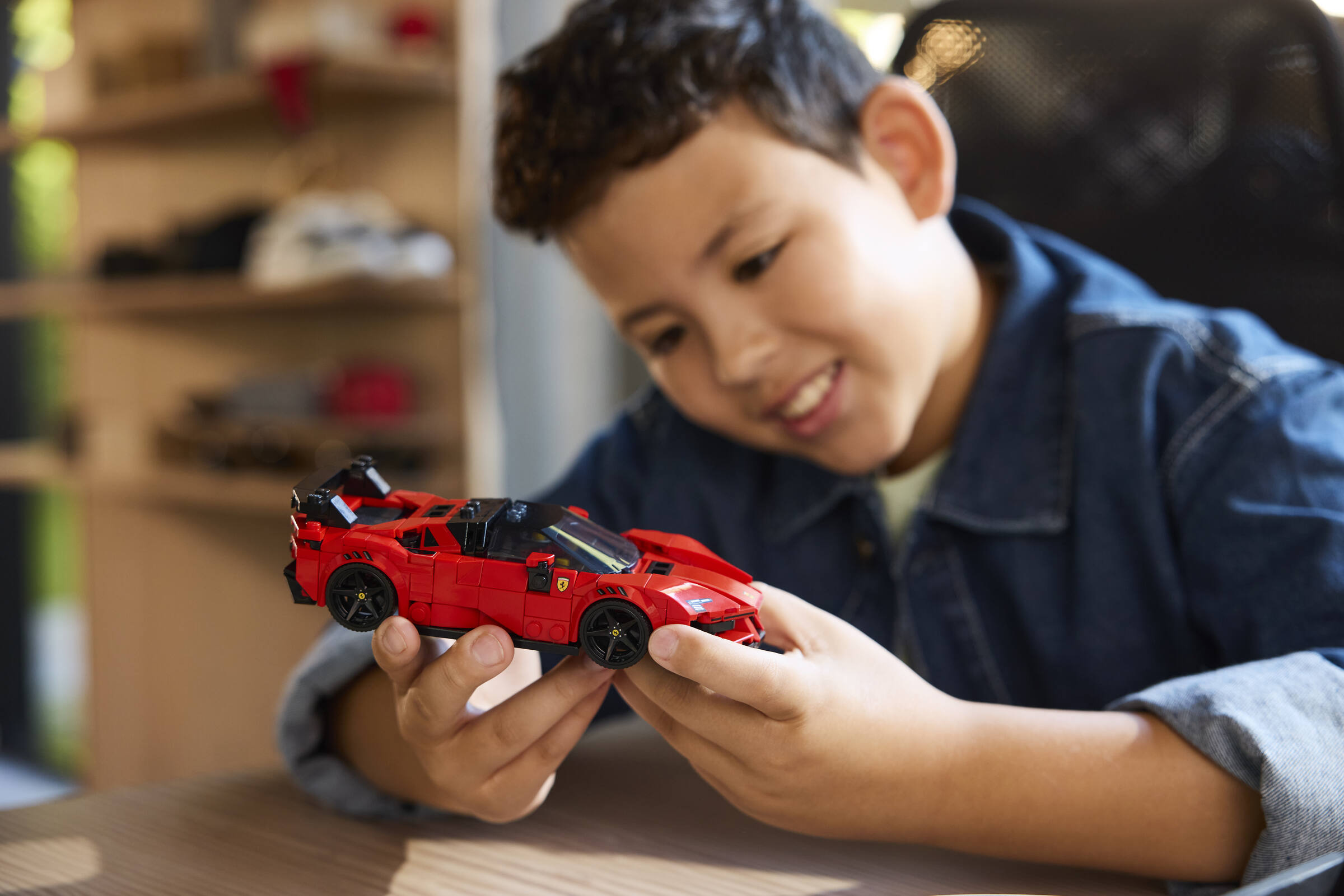 Ein Junge in einer blauen Jeansjacke lächelt und hält ein rotes Spielzeugauto aus Bausteinen.