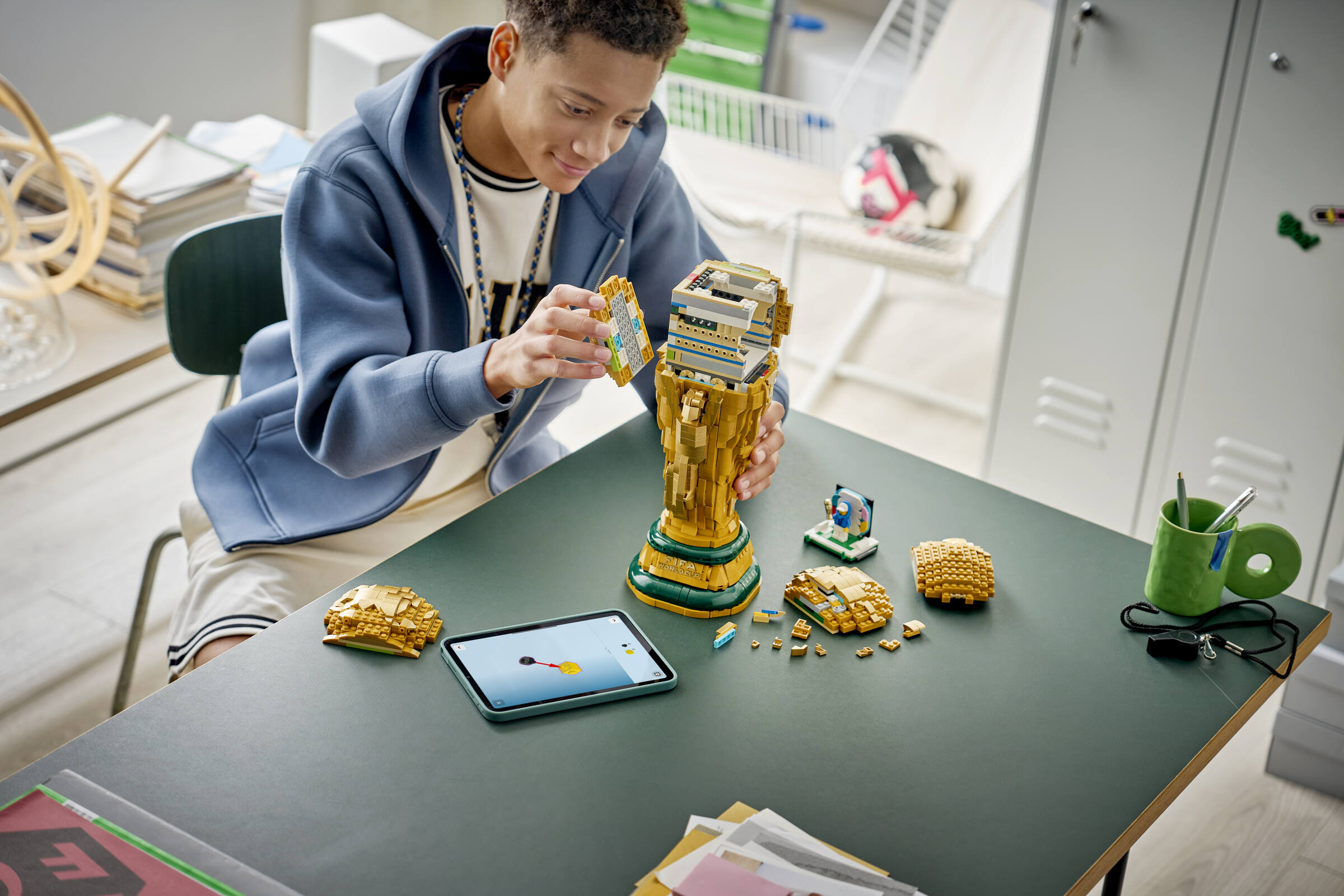 Ein Junge im blauen Hoodie baut eine große Lego-Trophäe an einem grünen Tisch.
