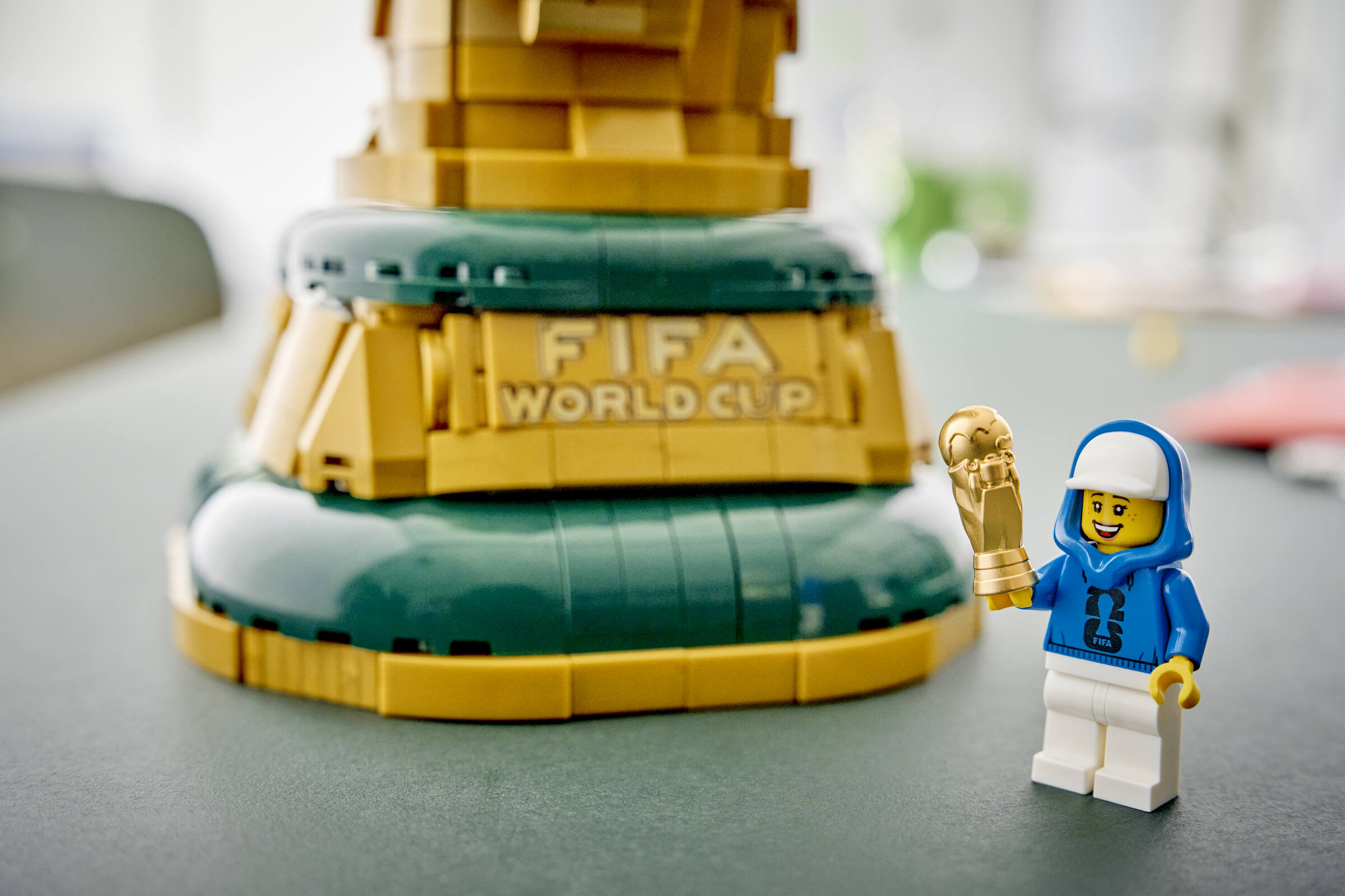 Eine Lego-Minifigur mit blauem Kapuzenpulli und weißer Mütze hält einen goldenen Pokal vor einem großen Lego-WM-Pokal.