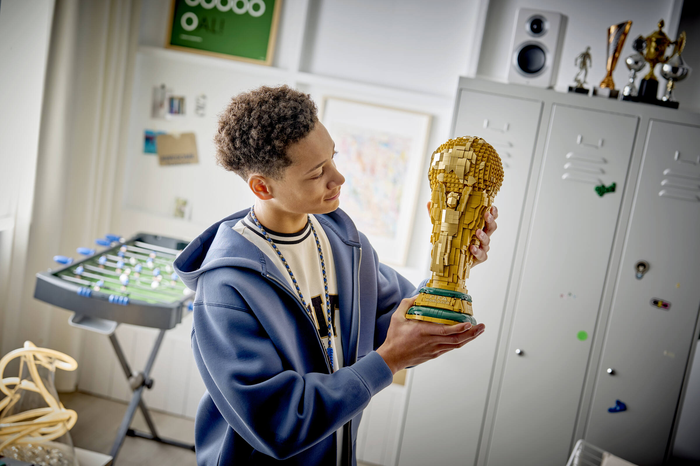 Ein junger Mann im blauen Hoodie hält stolz eine goldene LEGO FIFA WM-Trophäe in einem Zimmer.