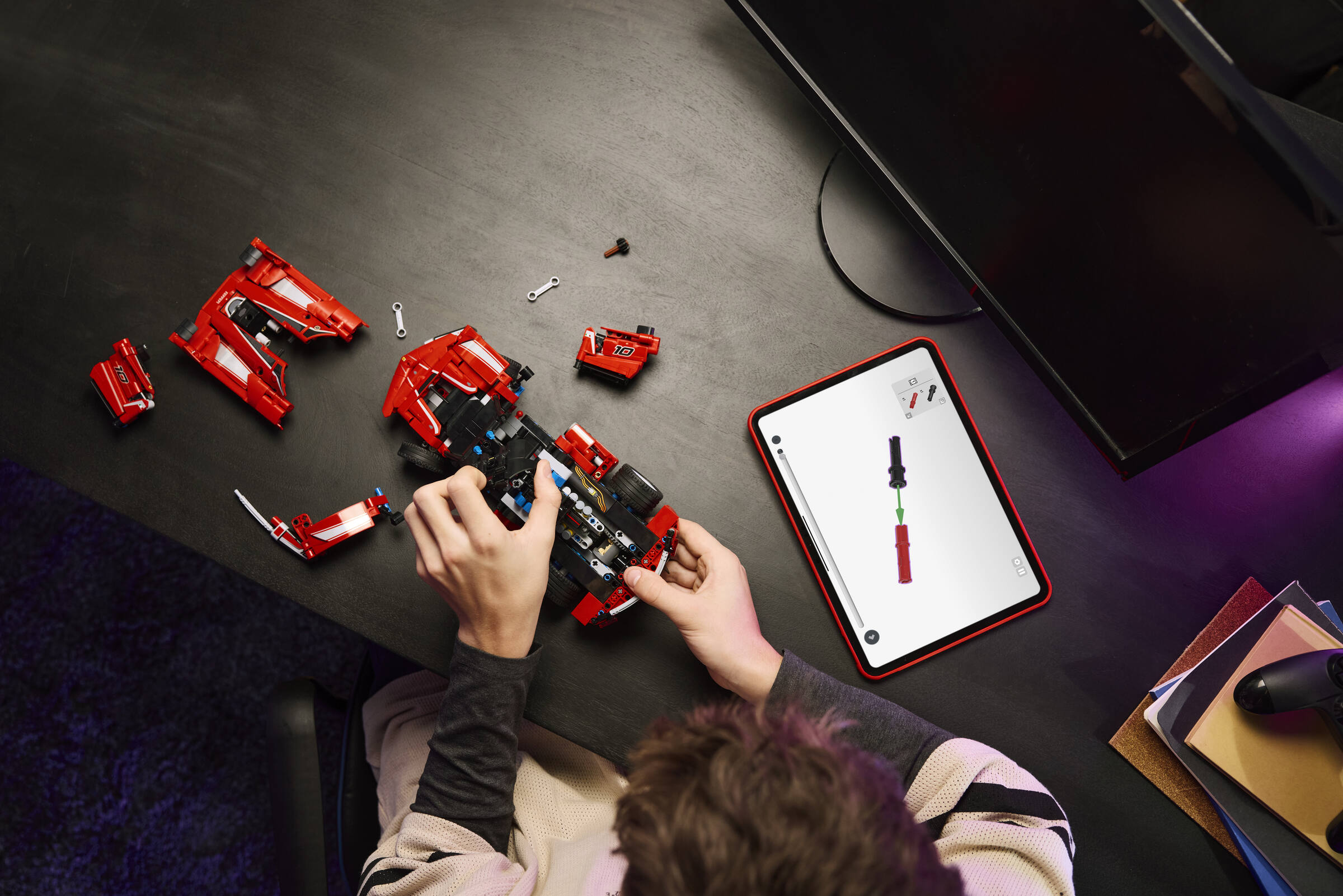 Une personne construit un modèle Lego rouge sur un bureau avec une tablette et un moniteur.