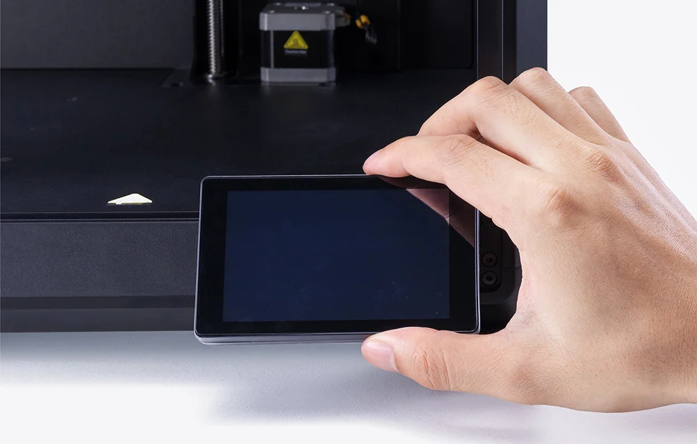 Egy kéz tartja a kis fekete LCD képernyőt egy 3D nyomtató előtt.