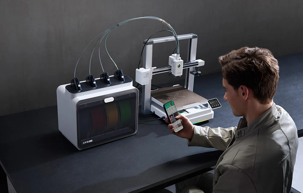 Egy férfi asztalnál ül, és okostelefonjával irányít egy izzószál-dobozos 3D nyomtatót.