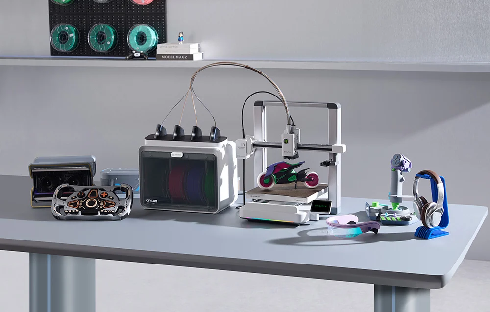 Egy 3D nyomtató izzószál dobozzal és színes kiegészítőkkel egy asztalon.