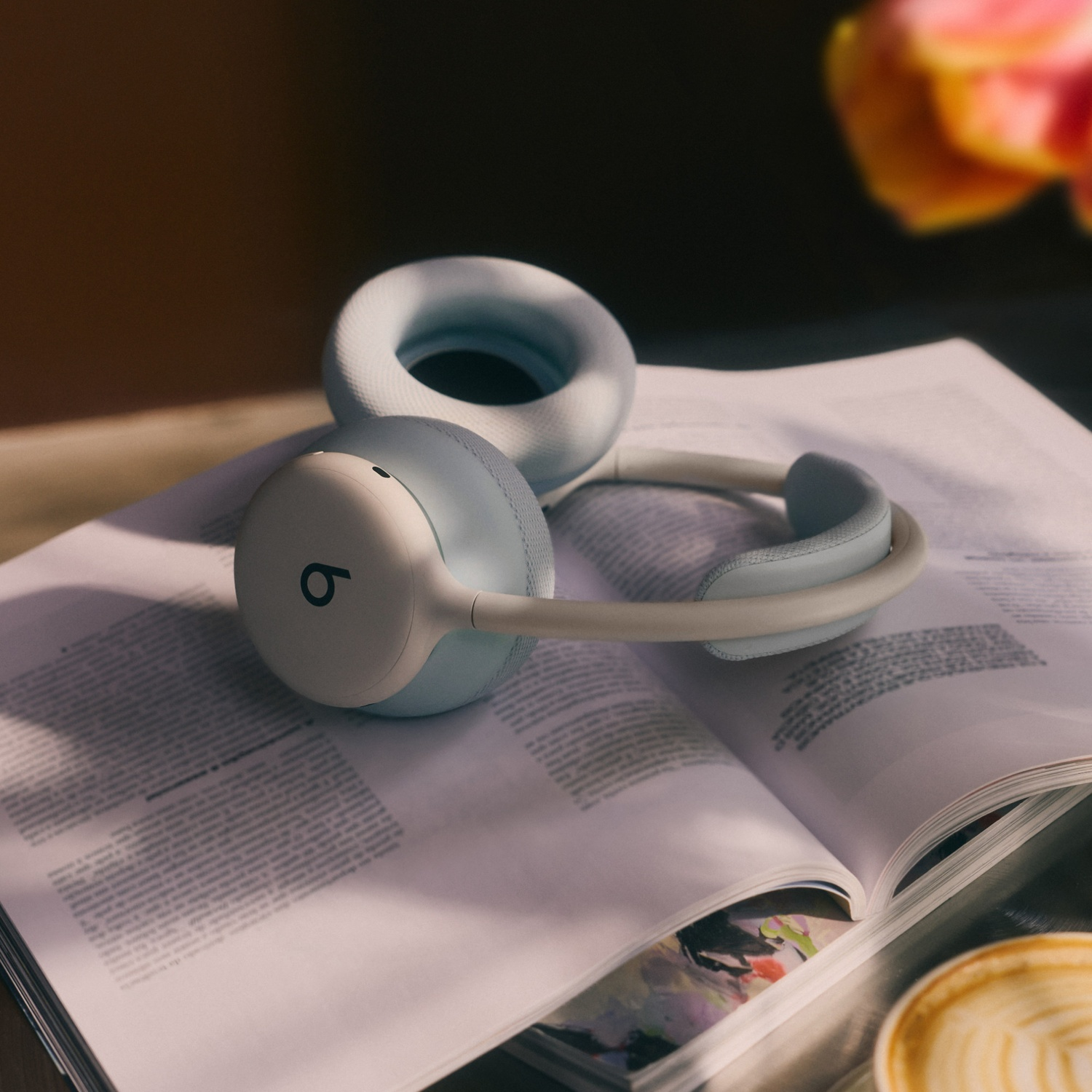Casque Beats bleu clair posé sur un magazine ouvert avec un café et une fleur en arrière-plan.
