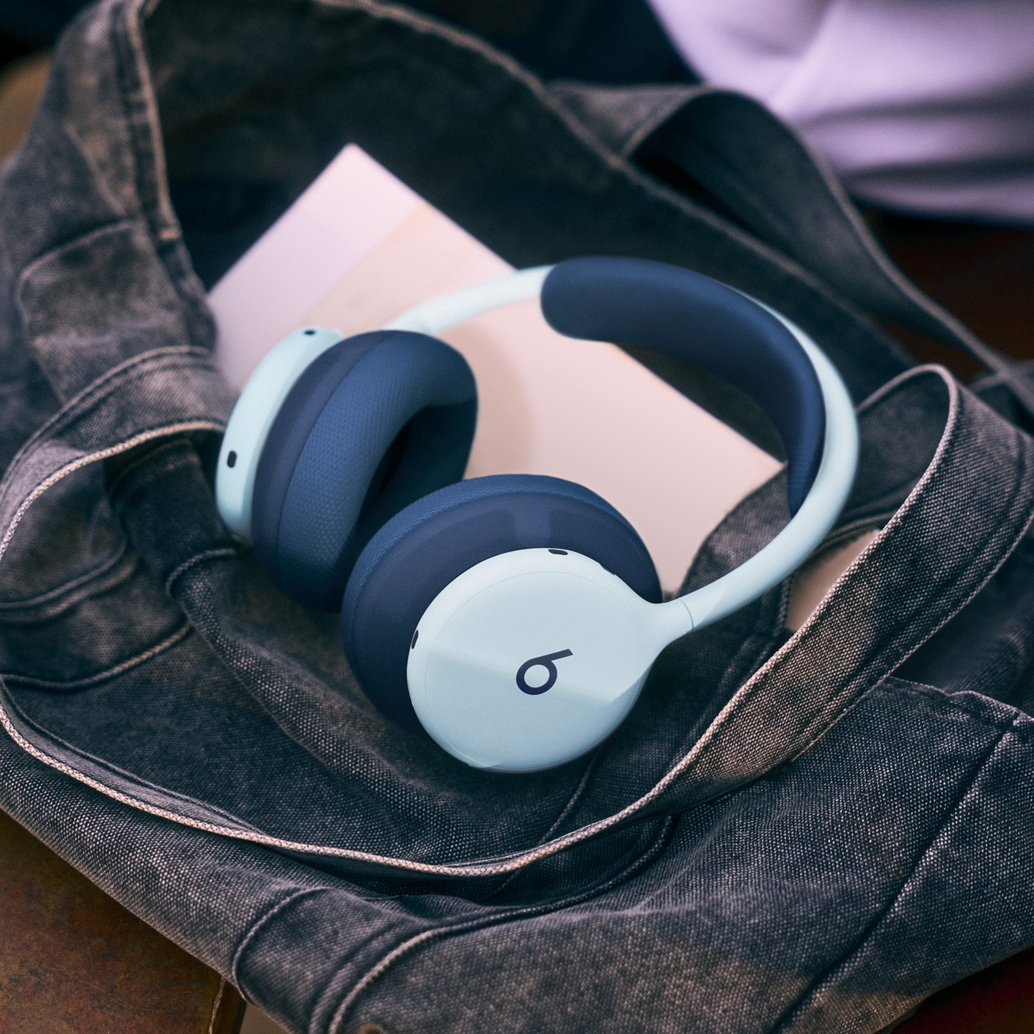 Casque supra-auriculaire sans fil bleu avec le logo Beats dans un sac cabas en denim sombre à côté d'un livre.