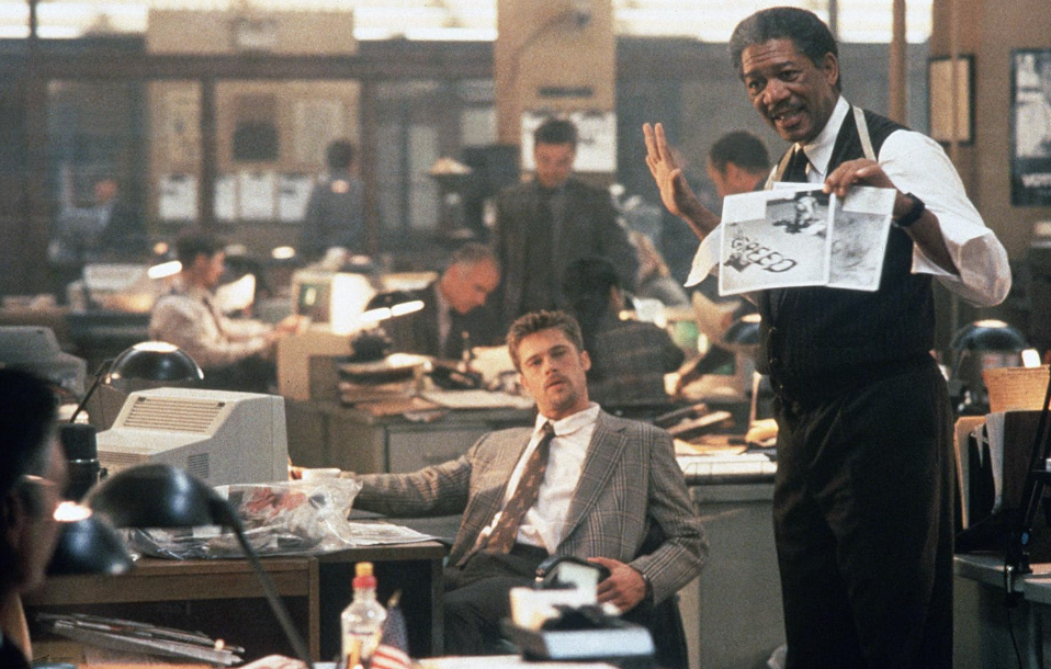 Morgan Freeman hält Dokumente in einem belebten Büro hoch, während Brad Pitt am Schreibtisch sitzt.