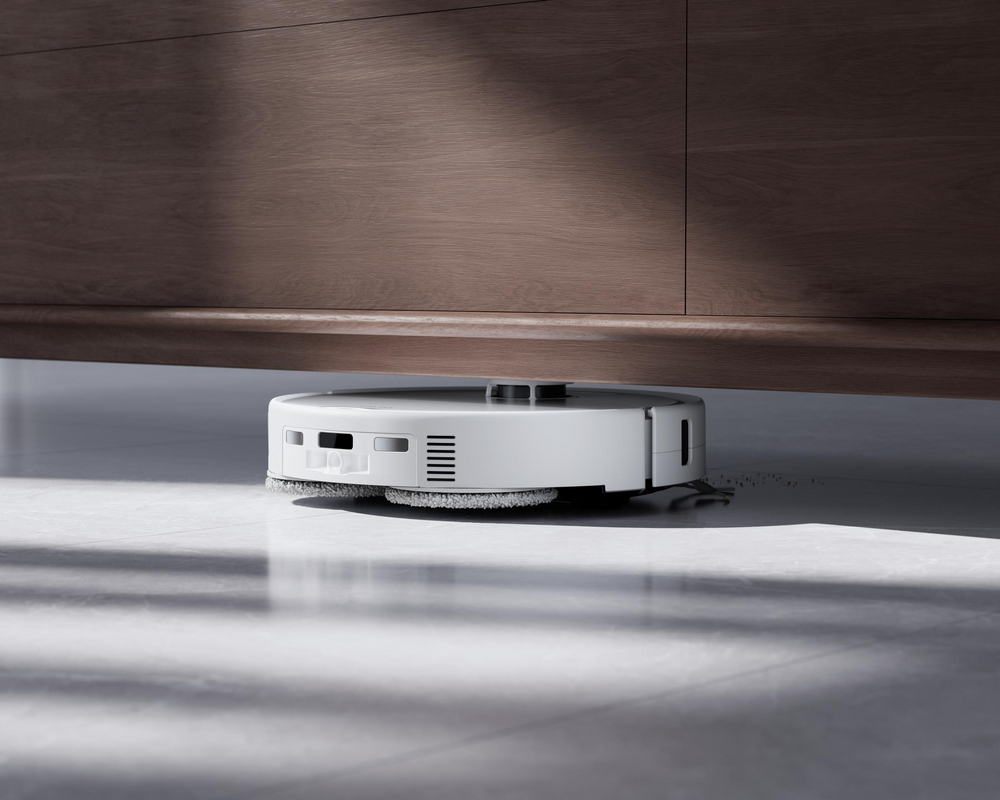Un robot aspirateur blanc nettoie le sol sous un meuble en bois.