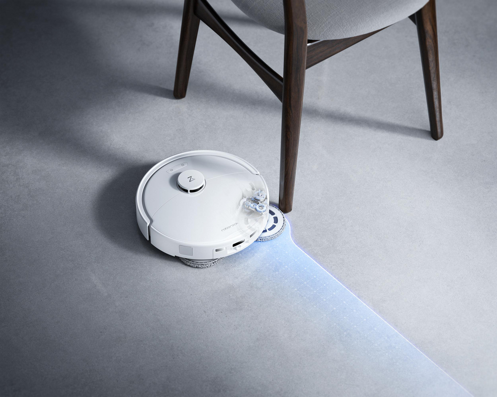 Un robot aspirateur blanc nettoie le sol en contournant un pied de chaise.