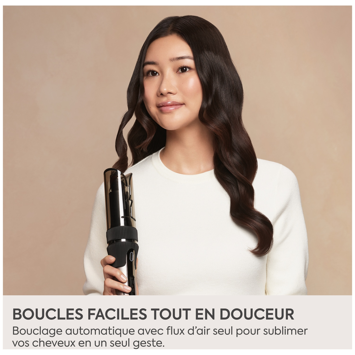 Une femme en pull blanc tient un fer à boucler noir sur fond neutre.