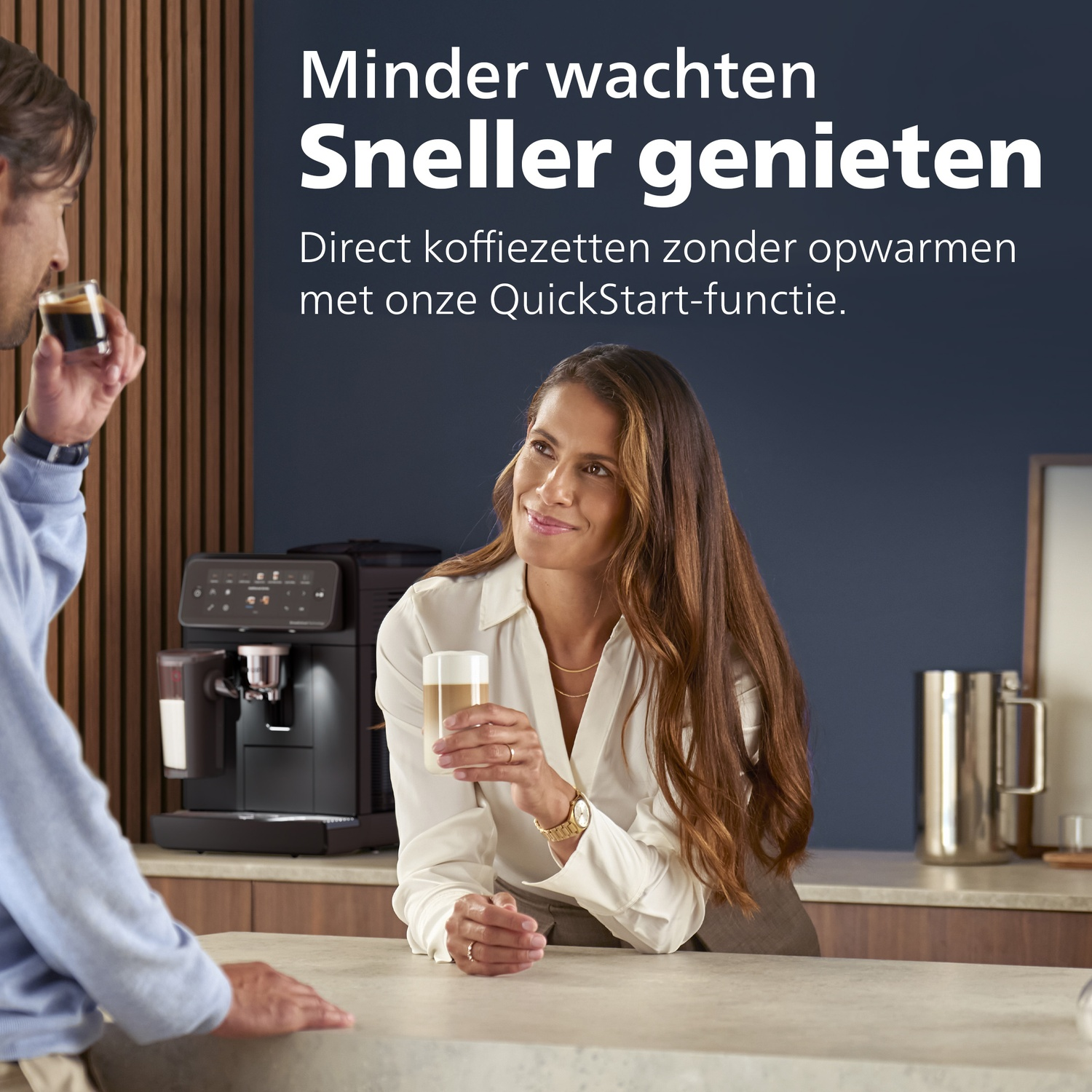 A woman holding a coffee cup looks at a man, with a coffee machine in the background.