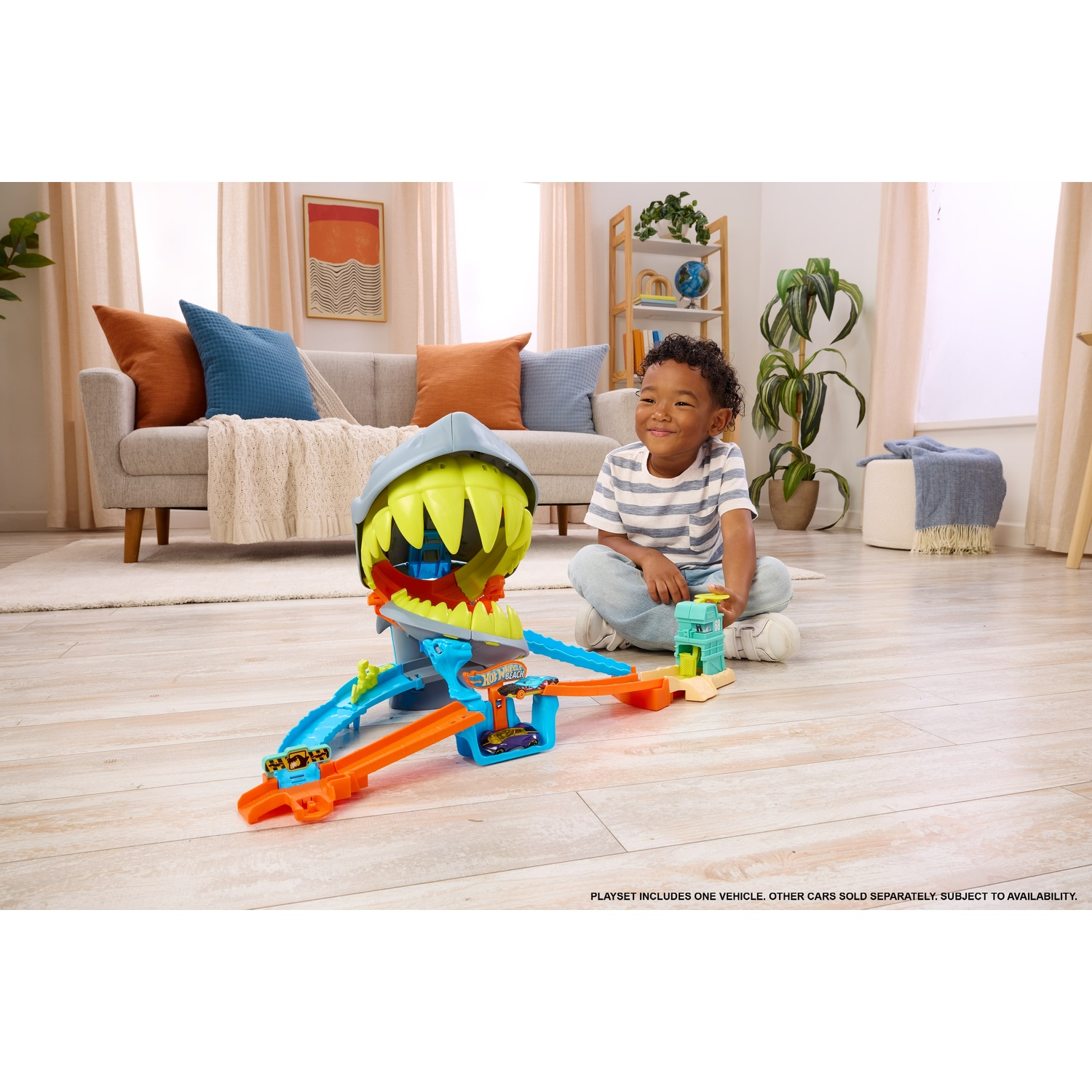 A young boy sits on a wooden living room floor playing with a colorful shark toy car track playset.