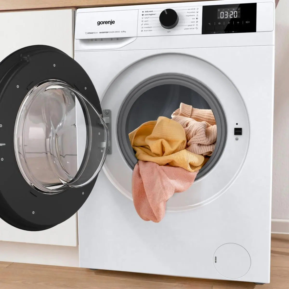 Fehér Gorenje mosógép nyitott ajtóval, sárga és rózsaszín ruhákkal a dobban.