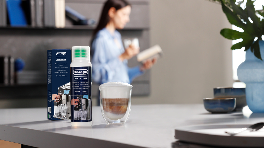 Eine De'Longhi Multiclean Flasche und Box stehen auf einer Küchentheke neben einem geschichteten Kaffee, eine Frau liest.