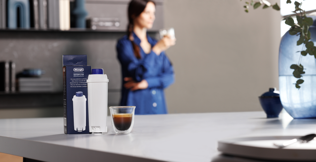 Ein De'Longhi-Wasserfilter, seine Verpackung und eine Tasse Espresso auf einer Theke, im Hintergrund eine Frau.