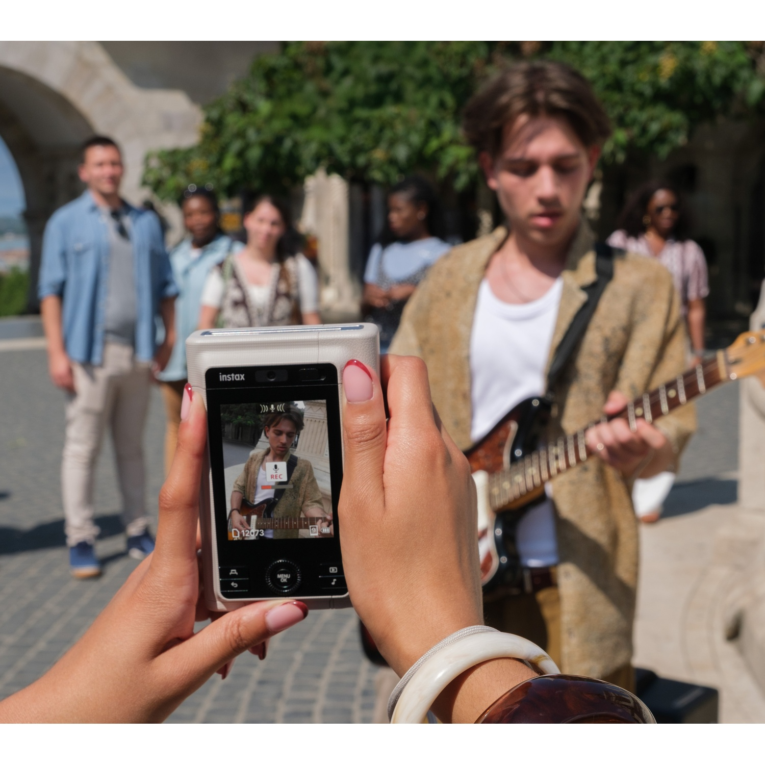 Une personne filme un jeune musicien de rue avec une guitare électrique à l'aide d'un appareil Instax, spectateurs.