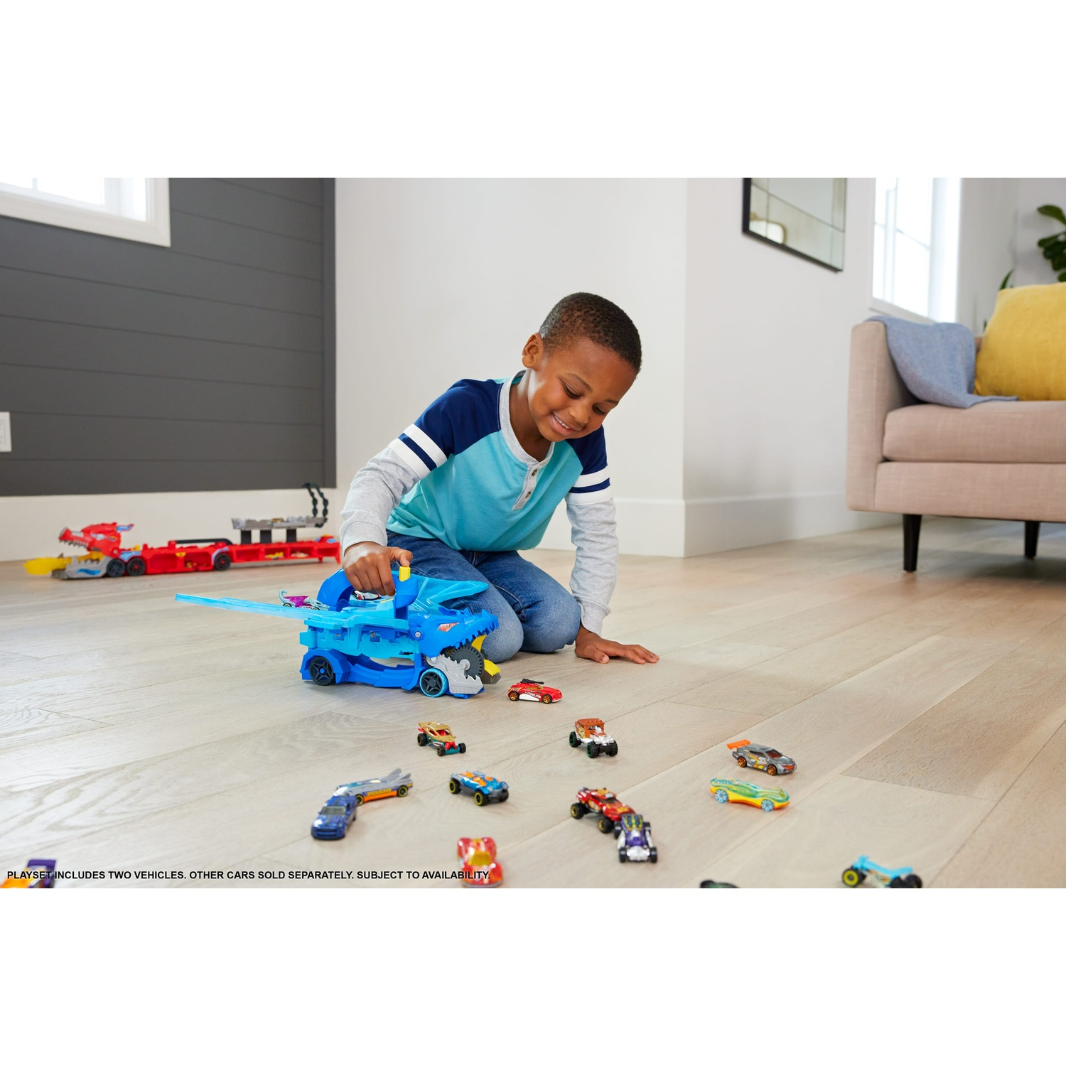 A young boy plays on a wood floor with a blue toy shark car carrier and many small toy cars.