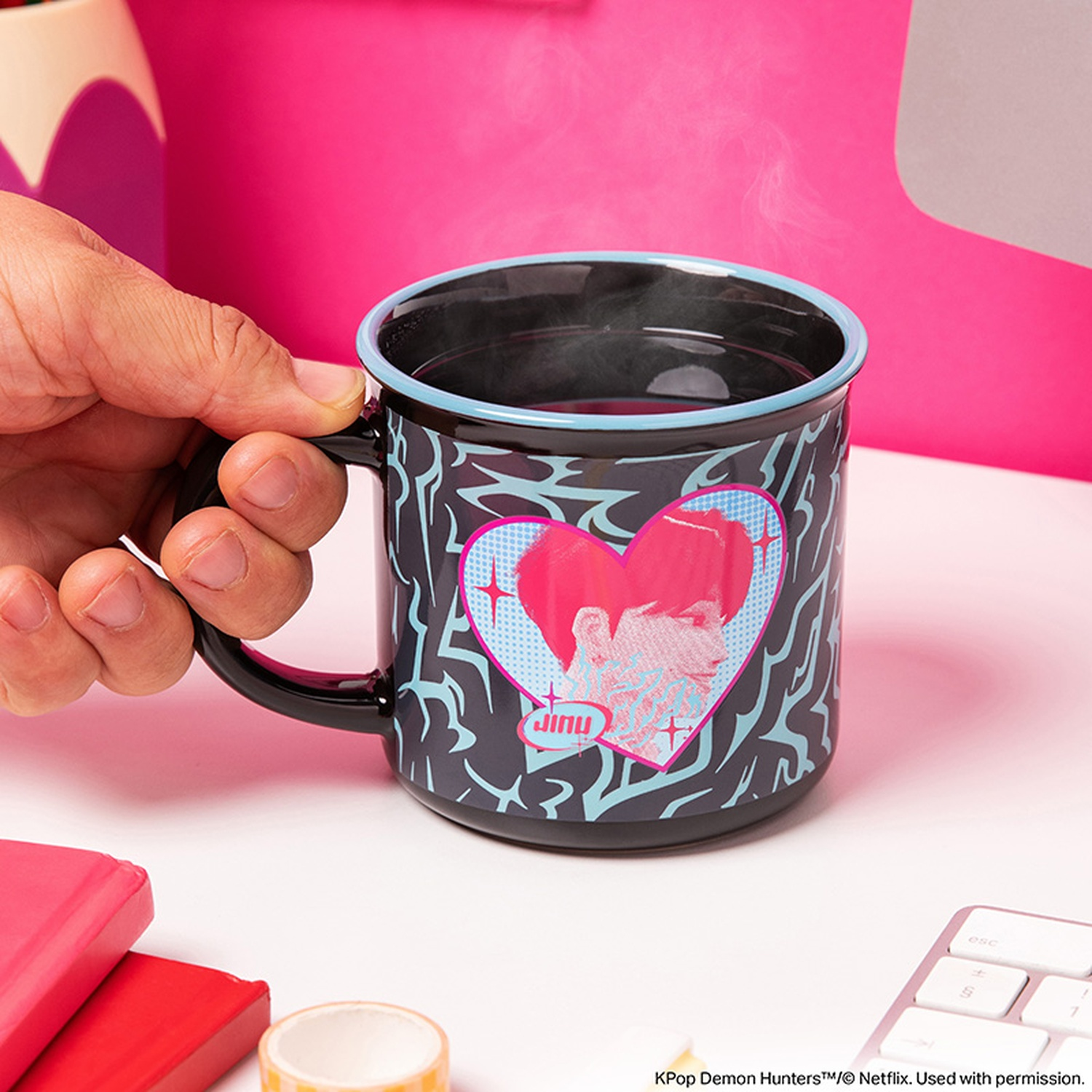 Une main tient une tasse noire avec un bord bleu et un cœur rose sur fond rose.