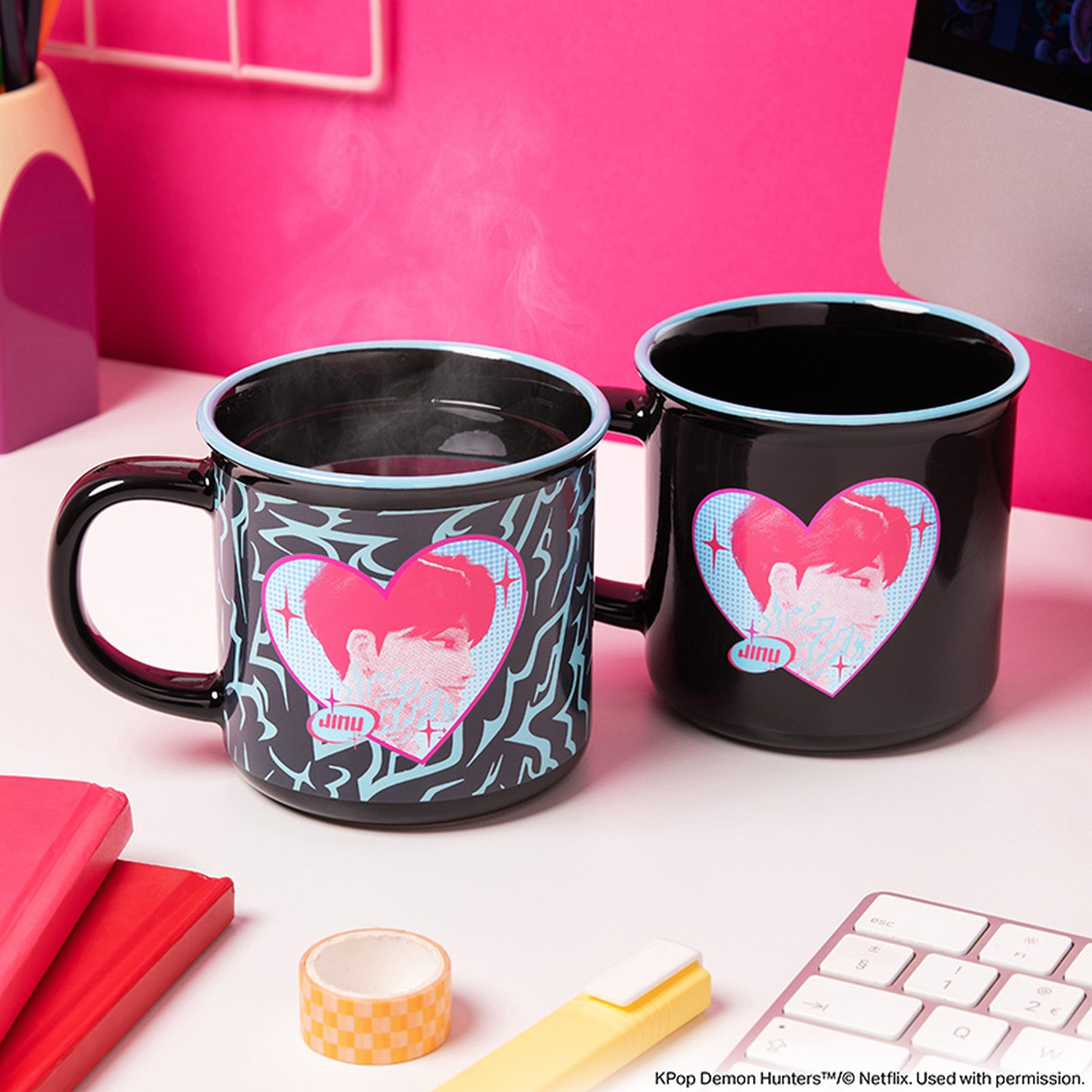 Deux tasses noires avec des cœurs et un graphisme de Jinu sur un bureau blanc avec fond rose.