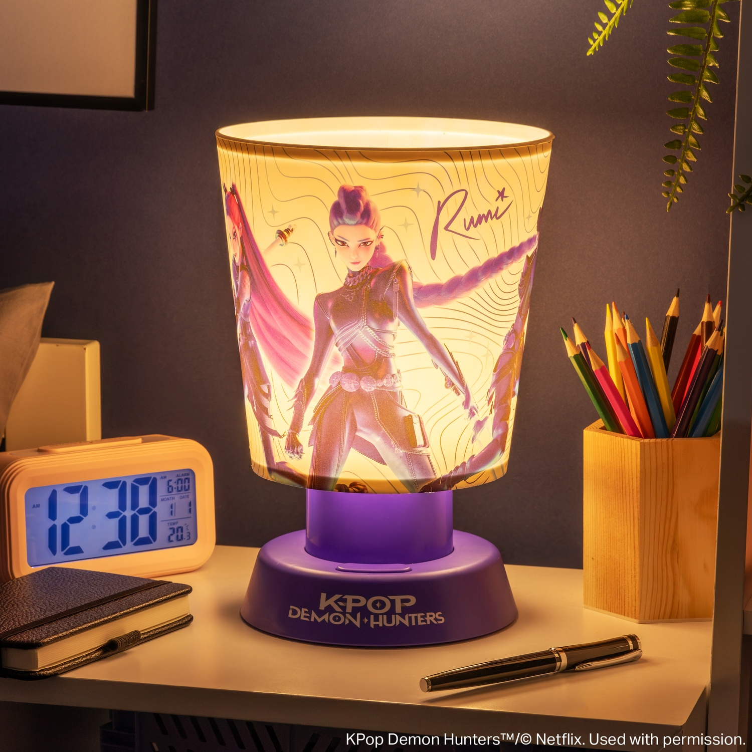 Une lampe K-Pop Demon Hunters allumée sur un bureau avec horloge numérique, carnet, stylo et crayons de couleur.
