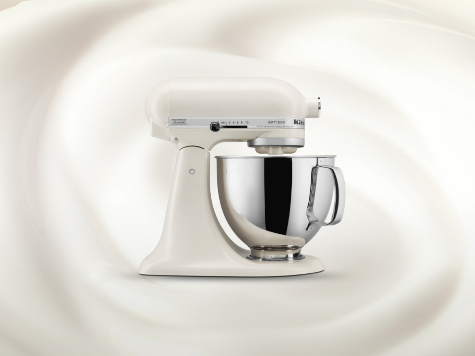 Cremefarbene KitchenAid Standmixer mit Edelstahlschüssel vor einem wirbelnden Hintergrund.