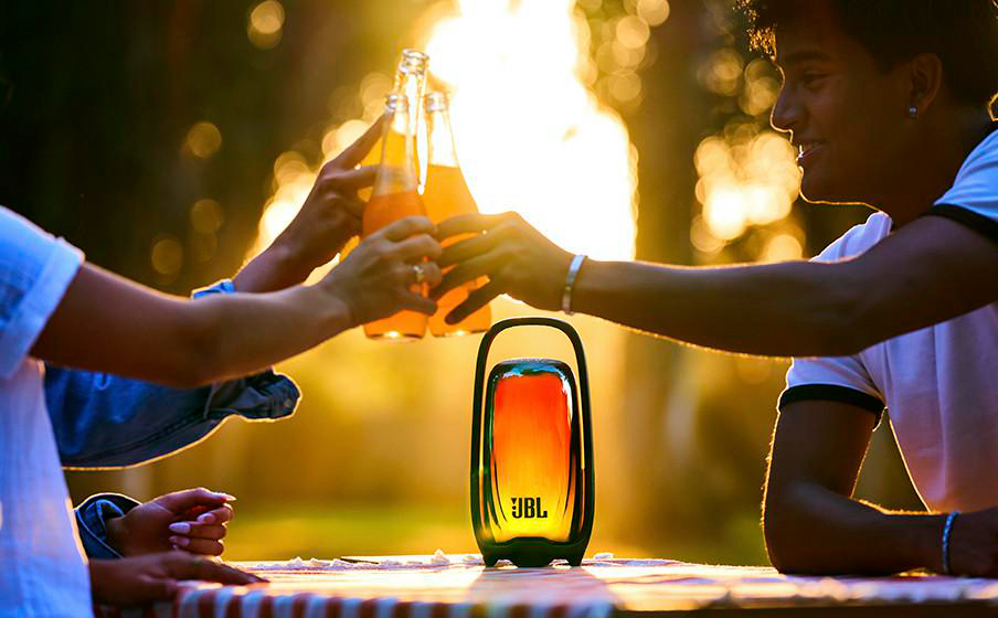 Freunde stoßen im Sonnenuntergang an einer Tisch an, auf dem ein beleuchteter JBL-Lautsprecher steht.