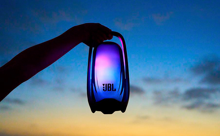 Eine Hand hält einen beleuchteten JBL-Lautsprecher vor einen blauen und gelben Abendhimmel.