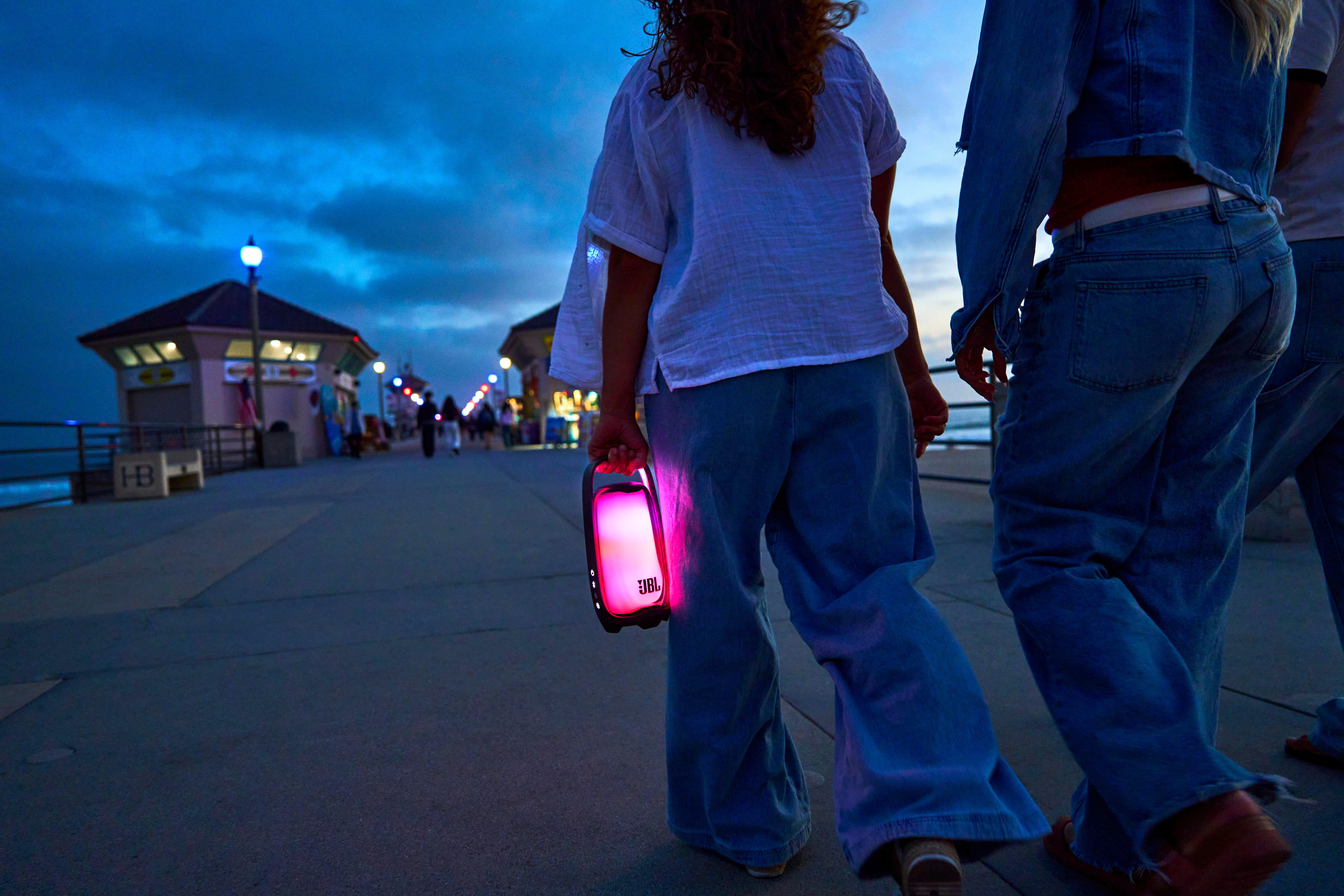 Personen gehen in der Dämmerung über einen Pier und tragen einen leuchtend rosa JBL-Lautsprecher.