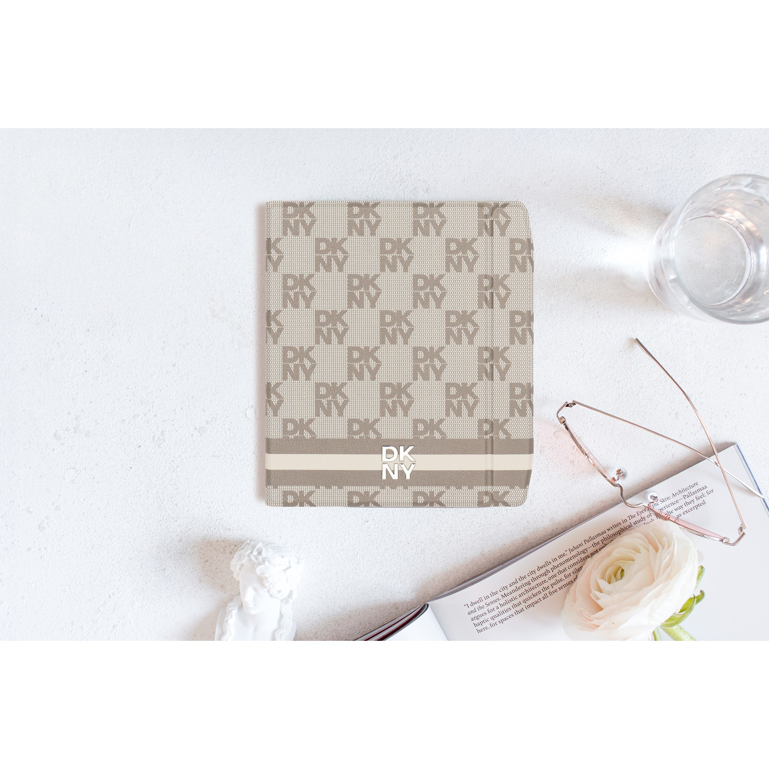 Een flat lay met een DKNY notitieboek, bril, witte roos, glas en beeldje op een witte ondergrond.
