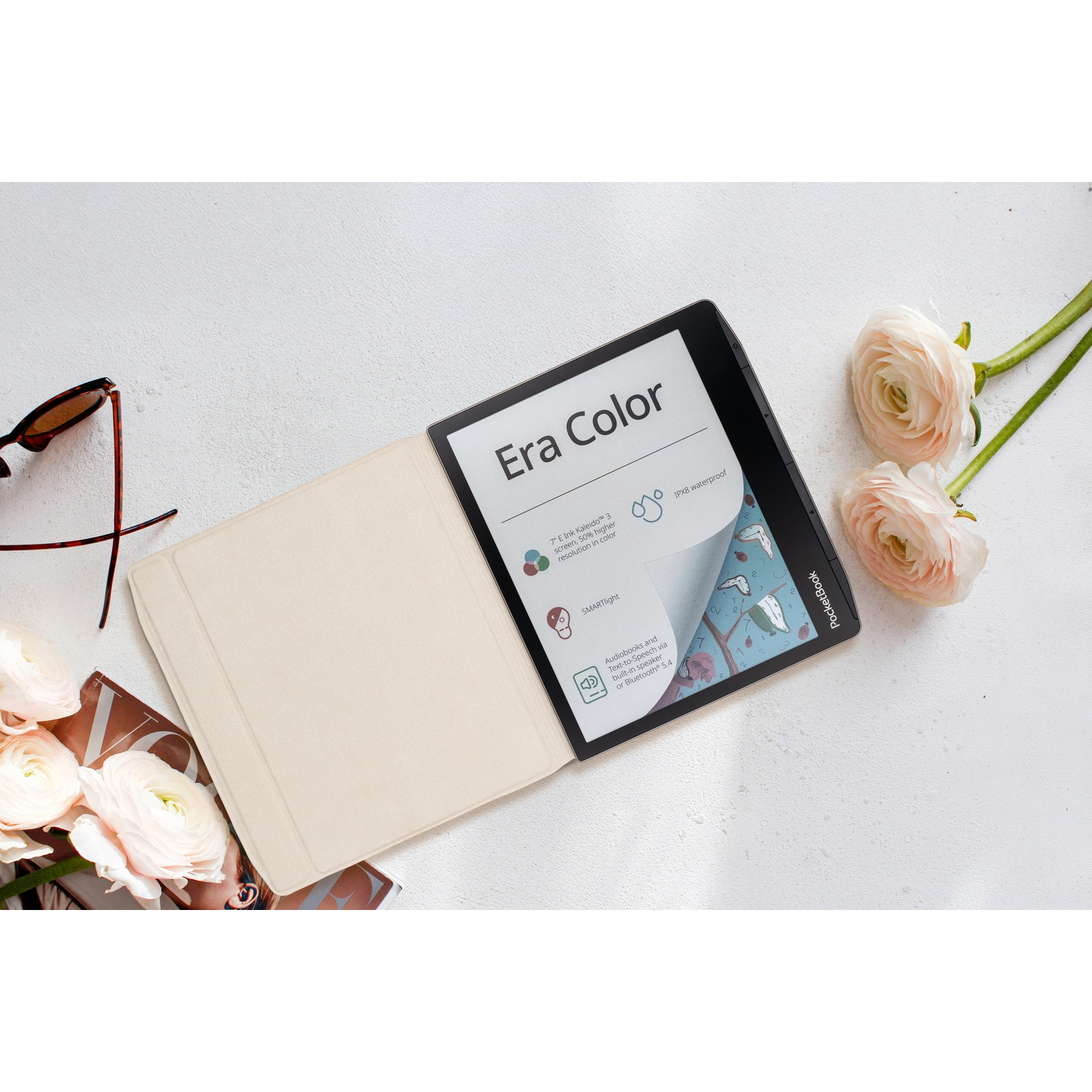 Een PocketBook Era Color e-reader op een witte tafel, omringd door roze bloemen en een zonnebril.